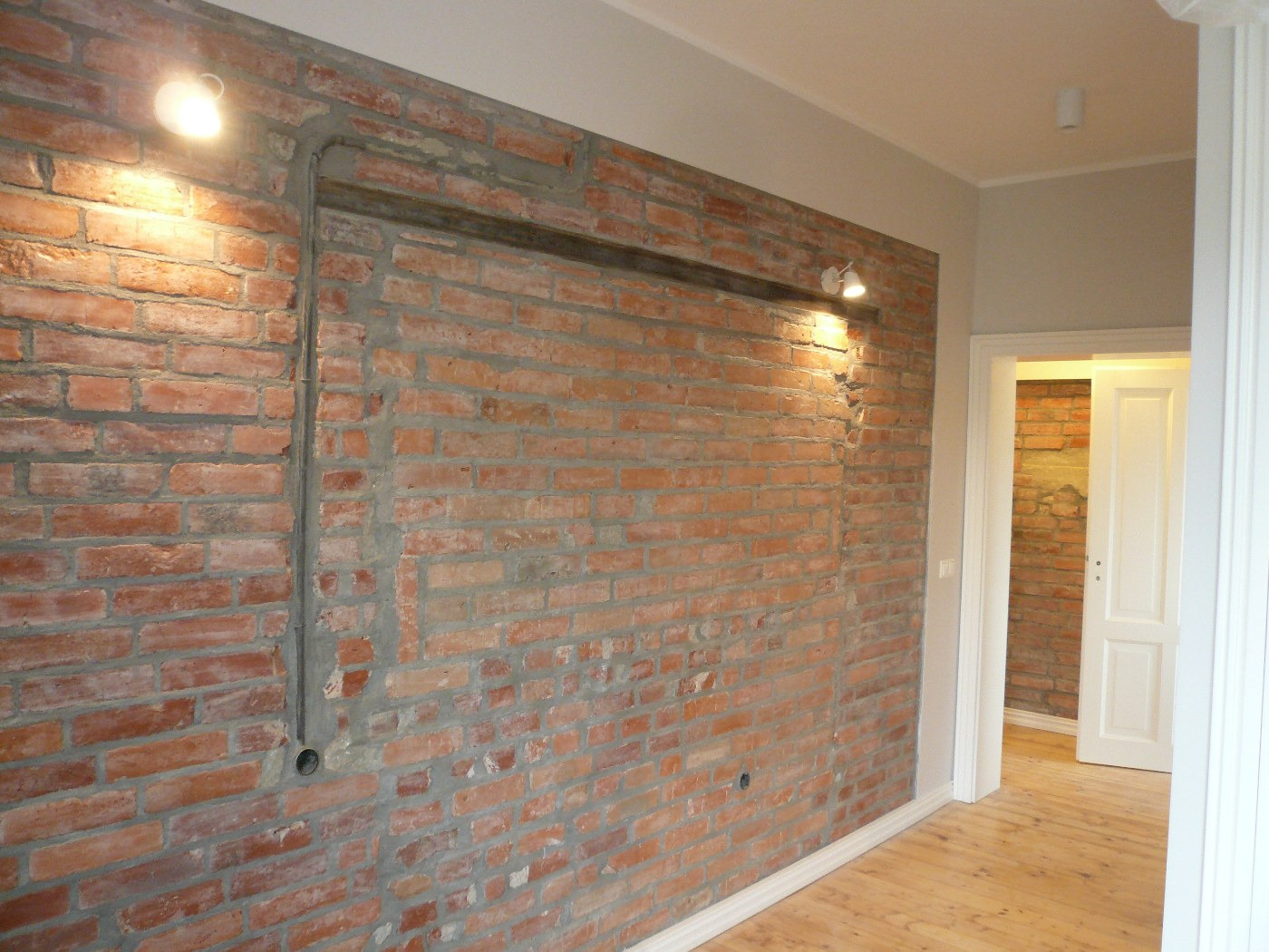 Ceglana ściana z widocznymi śladami po instalacjach elektrycznych i hydraulicznych, oświetlona dwoma kinkietami, z drewnianą podłogą i widokiem na kolejne pomieszczenie przez otwarte drzwi.