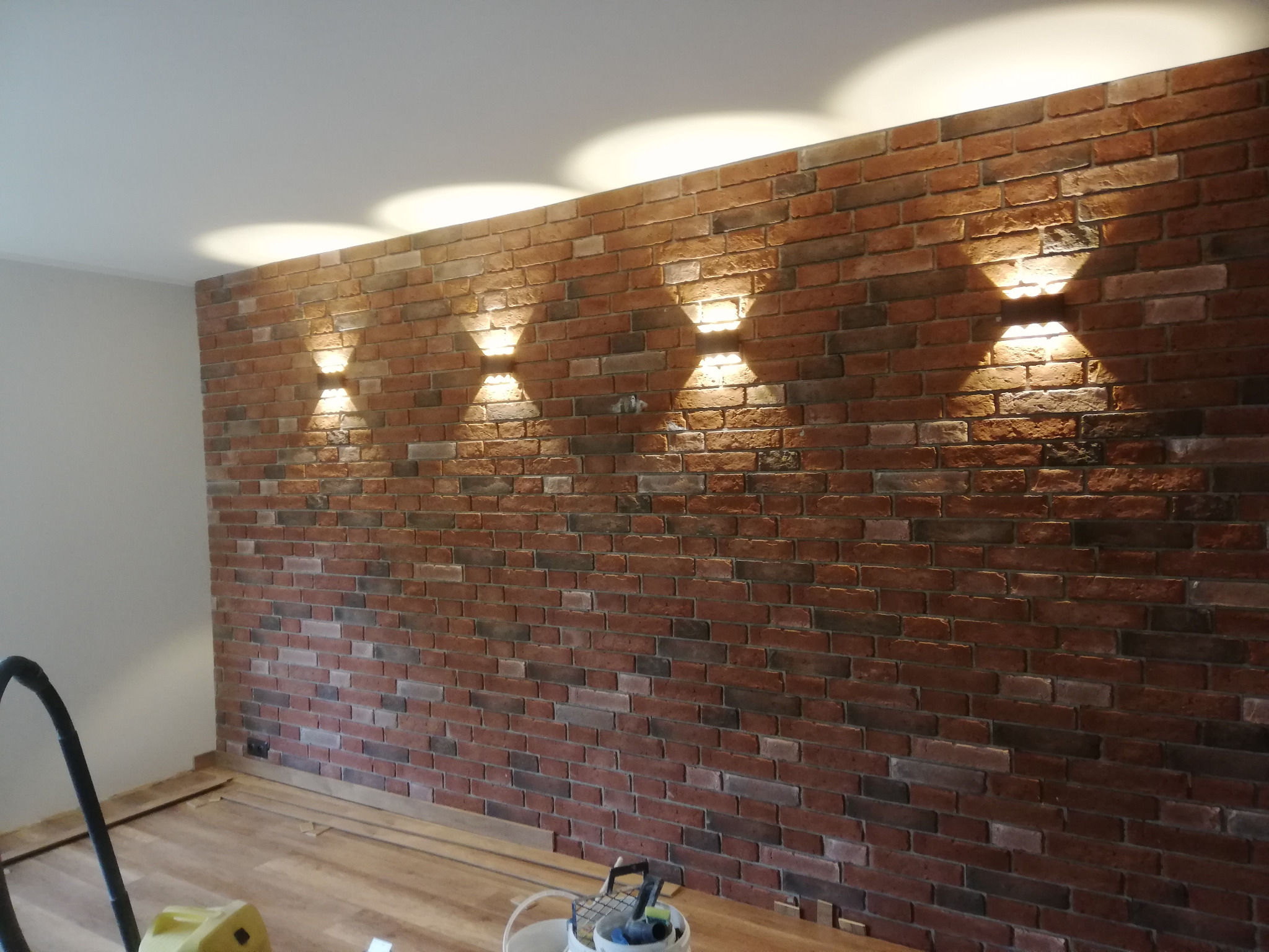 Ściana z cegły dekoracyjnej w pomieszczeniu z zamontowanymi lampami ściennymi, widoczne narzędzia i materiały na podłodze.