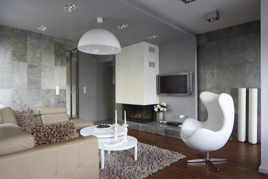 Nowoczesny salon z kominkiem obłożonym szarym kamieniem, beżowa skórzana sofa z dekoracyjnymi poduszkami, biały fotel Egg, dwa białe stoliki kawowe i telewizor na ścianie.
