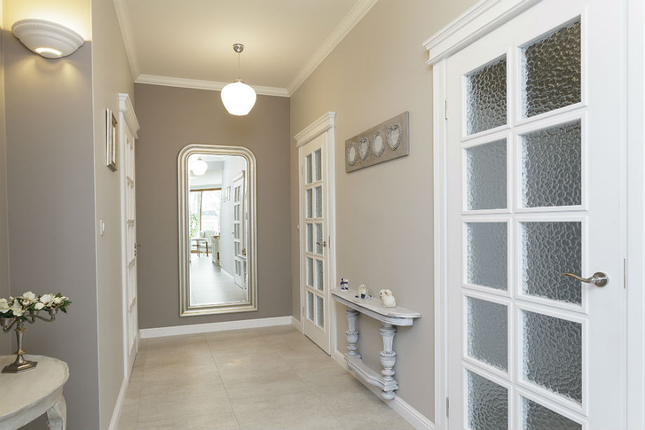 Jasny korytarz w Opolu z szarymi ścianami, białymi drzwiami z dekoracyjnym szkłem, dużym lustrem w srebrnej ramie, konsolą z figurkami i kwiatami, oświetlony lampą sufitową i kinkietami.