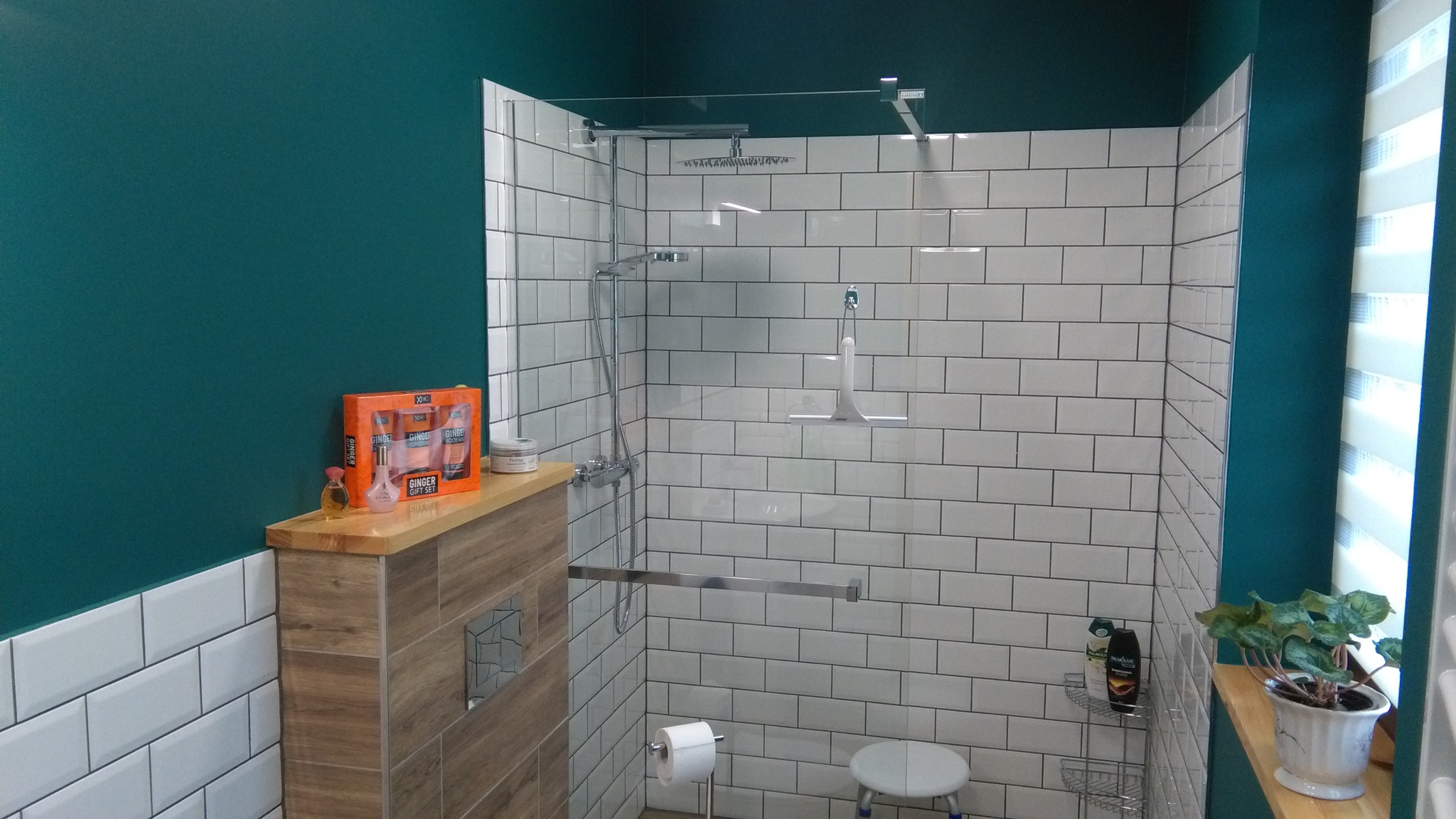 Nowoczesna kabina prysznicowa z białymi, prostokątnymi płytkami, szklaną ścianką i deszczownicą, uzupełniona o drewnopodobną zabudowę z zestawem kosmetyków i rośliną doniczkową, na tle ściany...