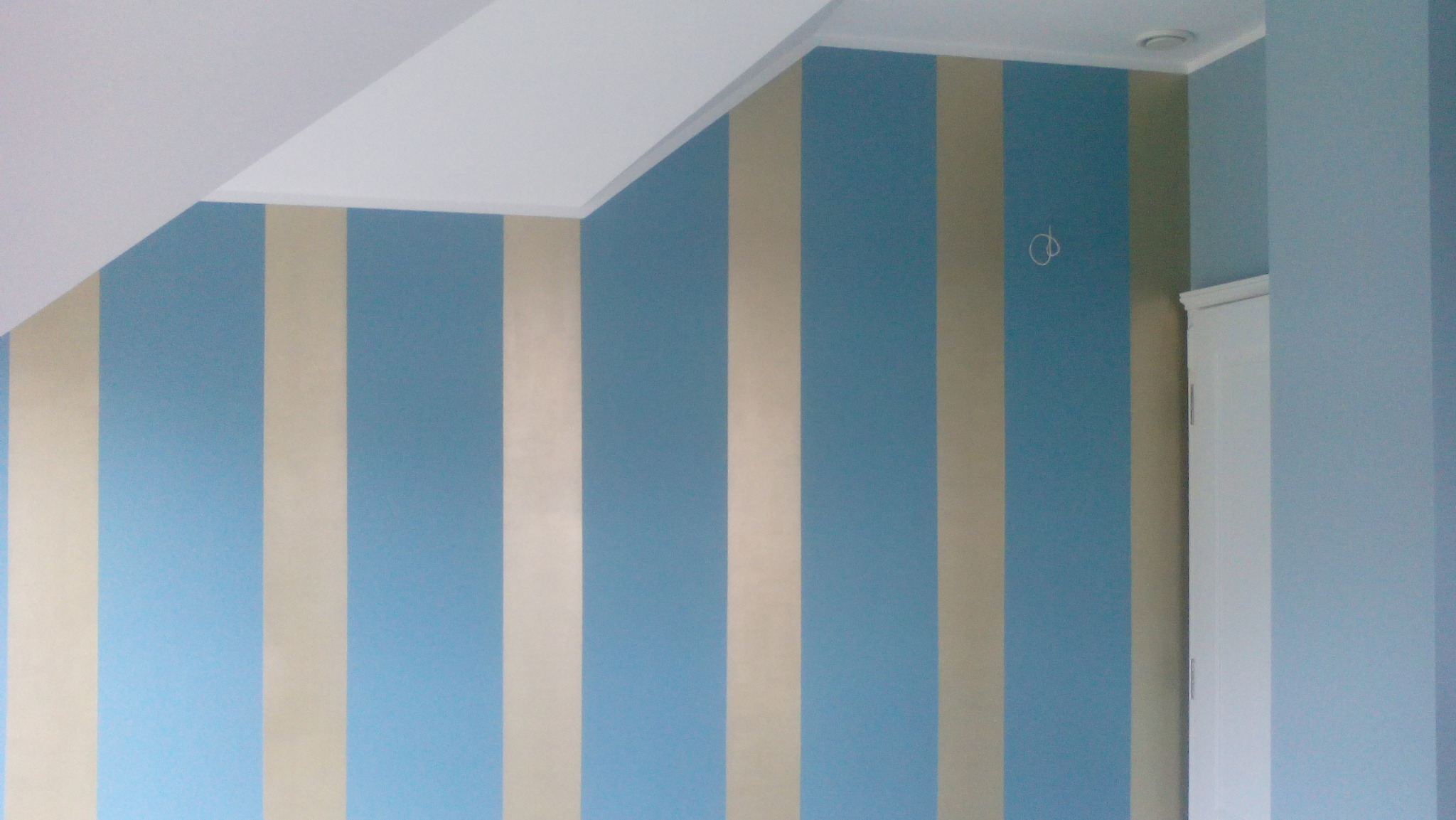 Pomalowana ściana w pionowe pasy koloru jasnoniebieskiego i beżowego, widoczny fragment sufitu pod skosem i biała listwa przysufitowa, oraz fragment ściany w jednolitym odcieniu niebieskiego...