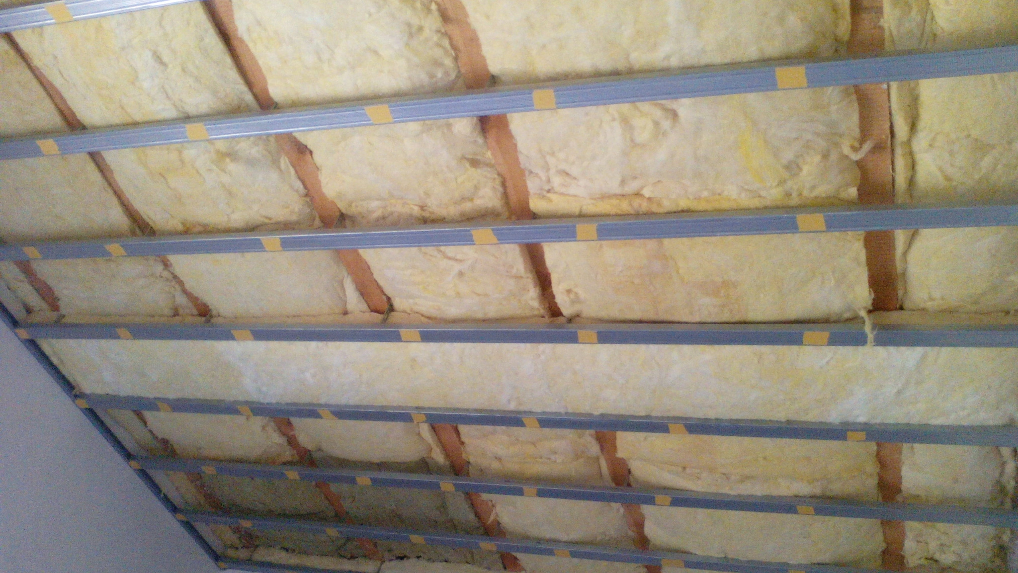 Poddasze z izolacją z wełny mineralnej, widoczne drewniane krokwie i metalowe profile mocujące, zabezpieczone żółtą taśmą.