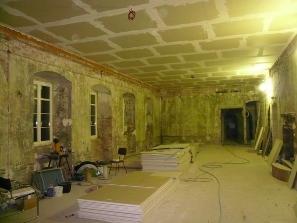 Przestronne wnętrze w trakcie prac wykończeniowych: sufit częściowo pokryty płytami gipsowo-kartonowymi, odsłonięte fragmenty ceglanej ściany z oknami, materiały budowlane i narzędzia na podłodze...