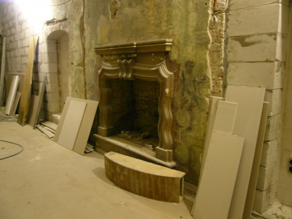 Wnętrze w trakcie prac z odsłoniętym kominkiem, widoczne płyty gipsowo-kartonowe i fragmenty starej ściany z dekoracjami.