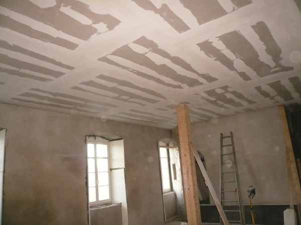 Wnętrze w trakcie prac przygotowawczych do wykończenia: surowa ściana, okno z białymi ramami, drewniane podpory i fragment sufitu z widocznymi łączeniami płyt i wstępnie nałożoną warstwą wyrównującą.