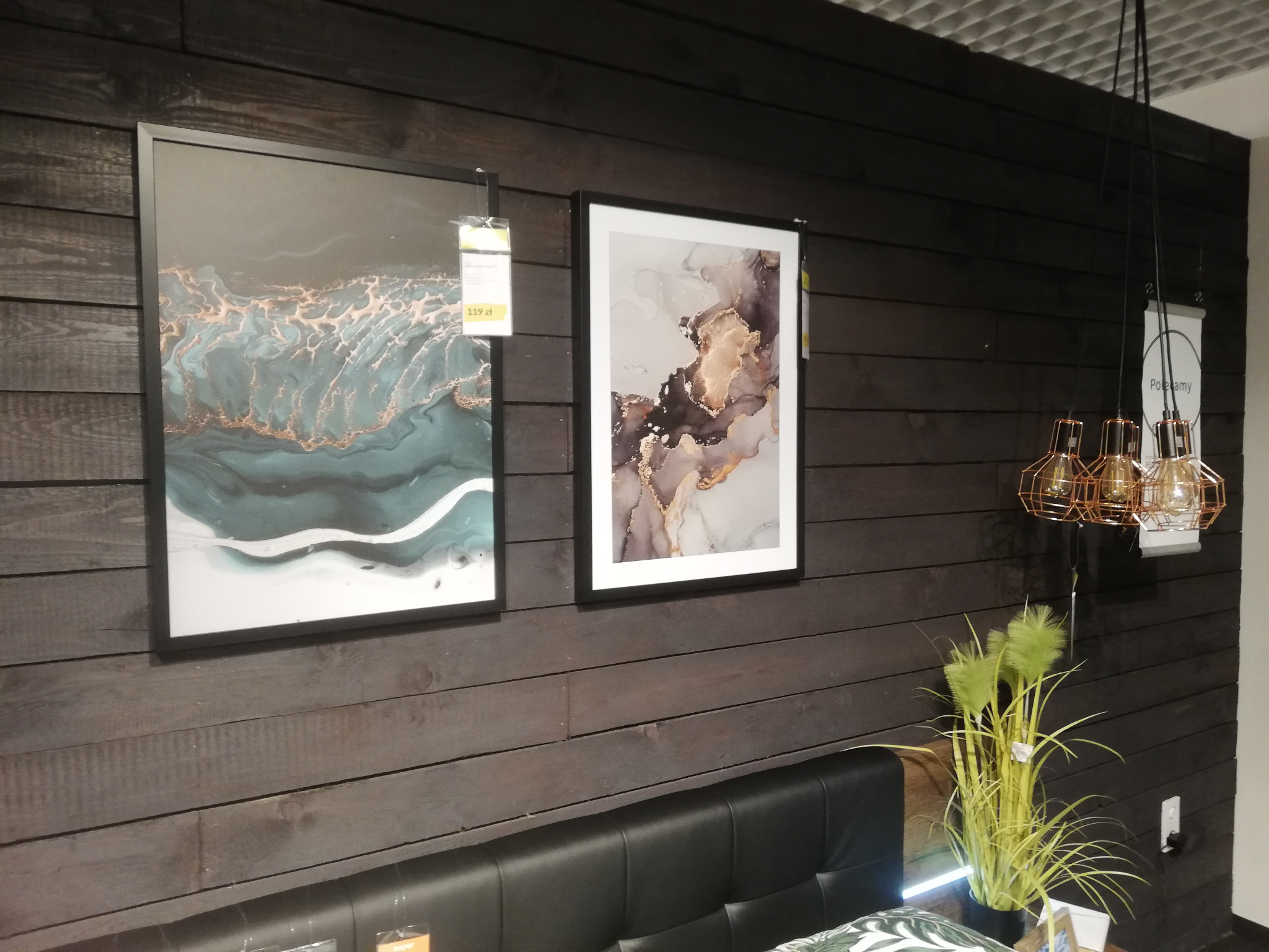 Ciemna drewniana ściana z dwoma obrazami w czarnych ramach, lampą wiszącą z miedzianymi kloszami i zieloną dekoracyjną trawą w doniczce, widoczny fragment wezgłowia łóżka.