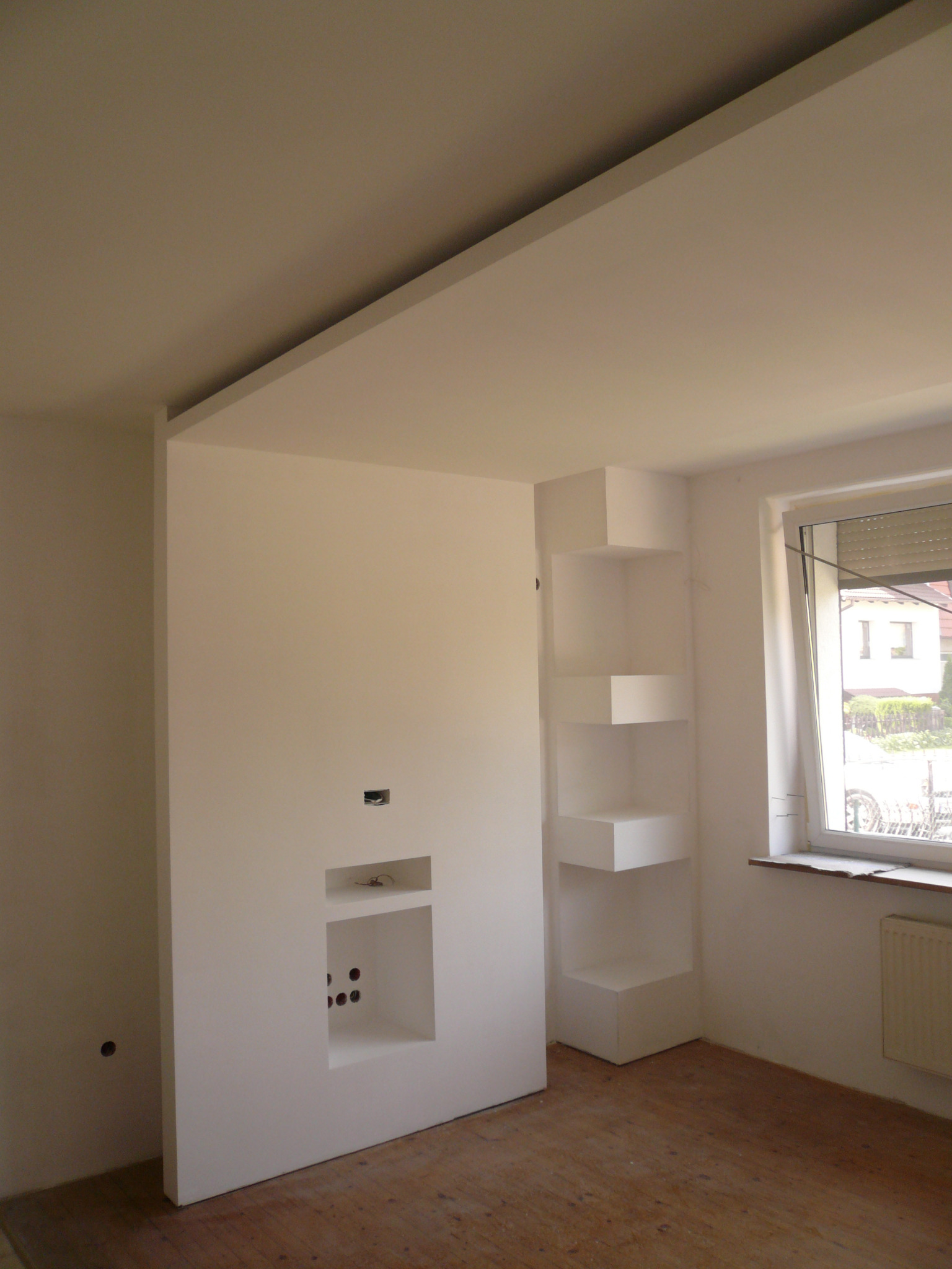 Wnętrze pokoju w trakcie prac: zabudowa z płyty gipsowo-kartonowej z wnękami na sprzęt RTV i półkami narożnymi, widoczne otwory na przewody, surowa podłoga, okno z żaluzjami.