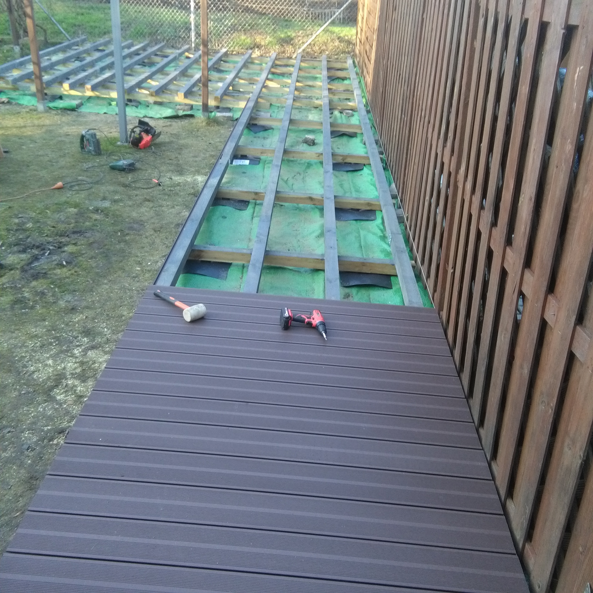 Montaż deski kompozytowej na przygotowanym ruszcie tarasu przy drewnianym ogrodzeniu, widoczny młotek gumowy i wkrętarka akumulatorowa.