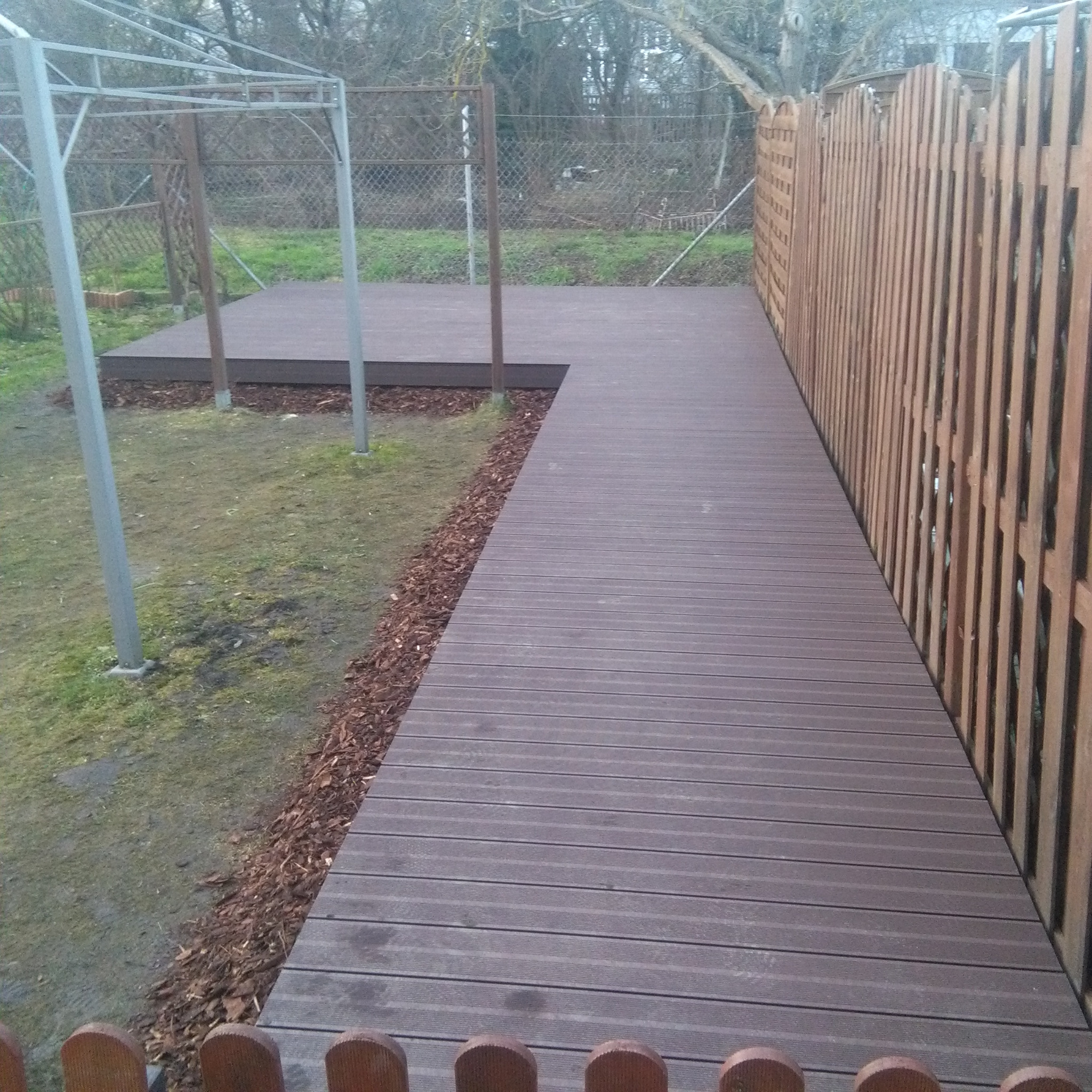 Kompozytowa deska tarasowa w kolorze ciemnego brązu, tworząca ścieżkę i podest w ogrodzie, z widocznym ogrodzeniem z drewnianych sztachet i metalową konstrukcją altany w tle.