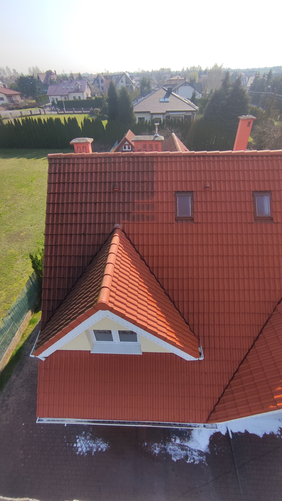 Czerwony dach z dachówki ceramicznej widziany z góry, z widocznym fragmentem elewacji i oknem dachowym, na tle zieleni i zabudowań.