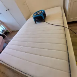 Materac z widocznym urządzeniem do czyszczenia, prawdopodobnie ekstrakcyjnego, z niebieskimi elementami, umieszczonym na jego powierzchni.