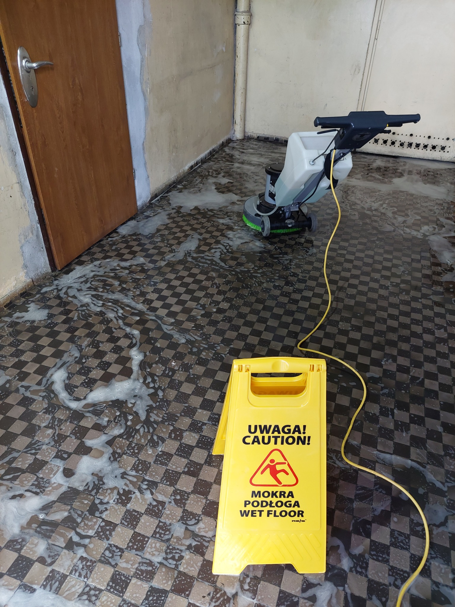 Automat do mycia podłóg w trakcie pracy na podłodze w szachownicę, widoczny żółty znak ostrzegawczy 'Uwaga! Mokra podłoga'.