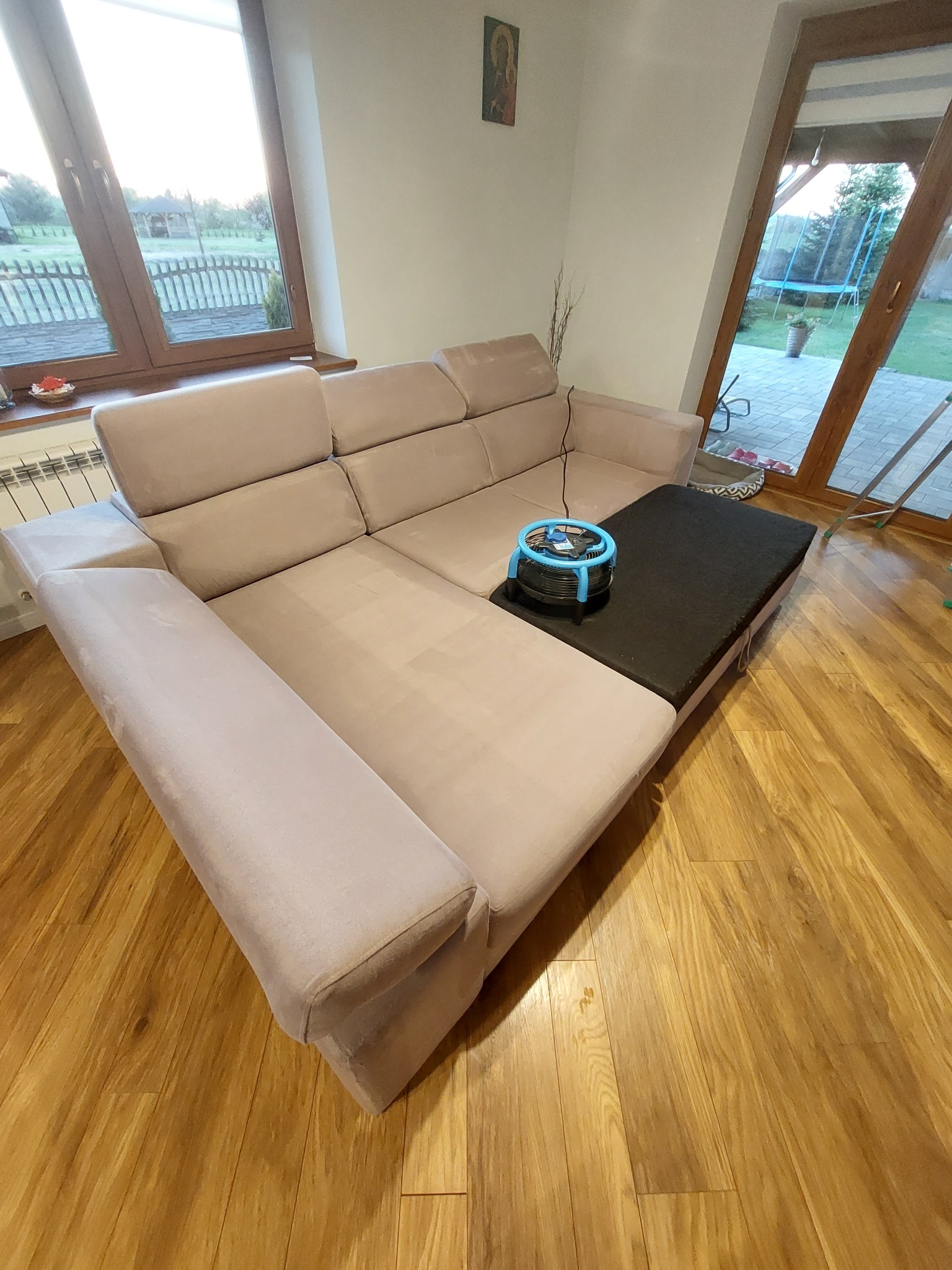Beżowa, modułowa kanapa z czarnym elementem i niebieskim wentylatorem na jasnym parkiecie, widok z góry, w tle okno z widokiem na ogród.