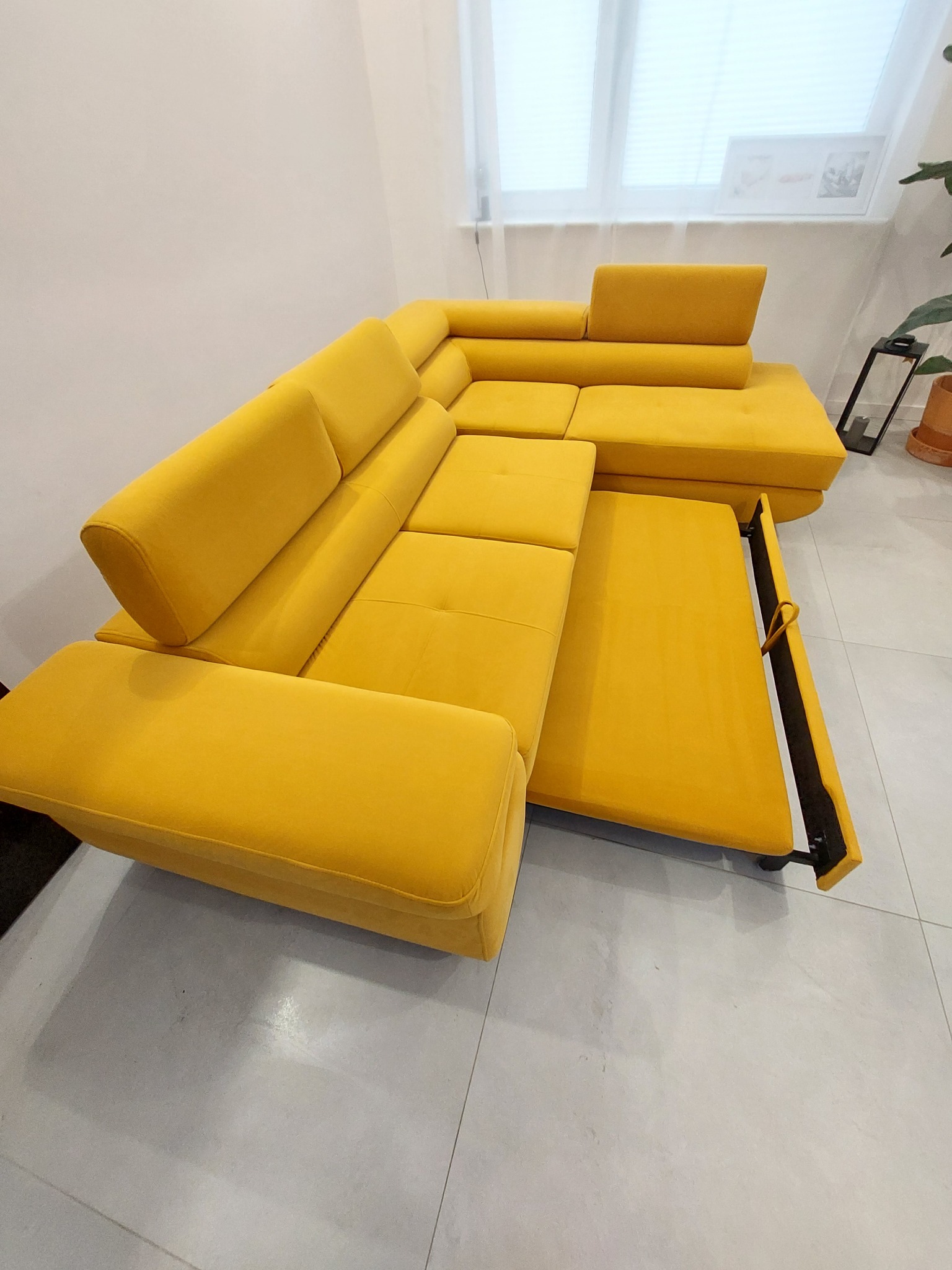 Żółta, rozkładana sofa narożna z wysuniętym elementem do spania, na tle jasnych ścian i płytek podłogowych.