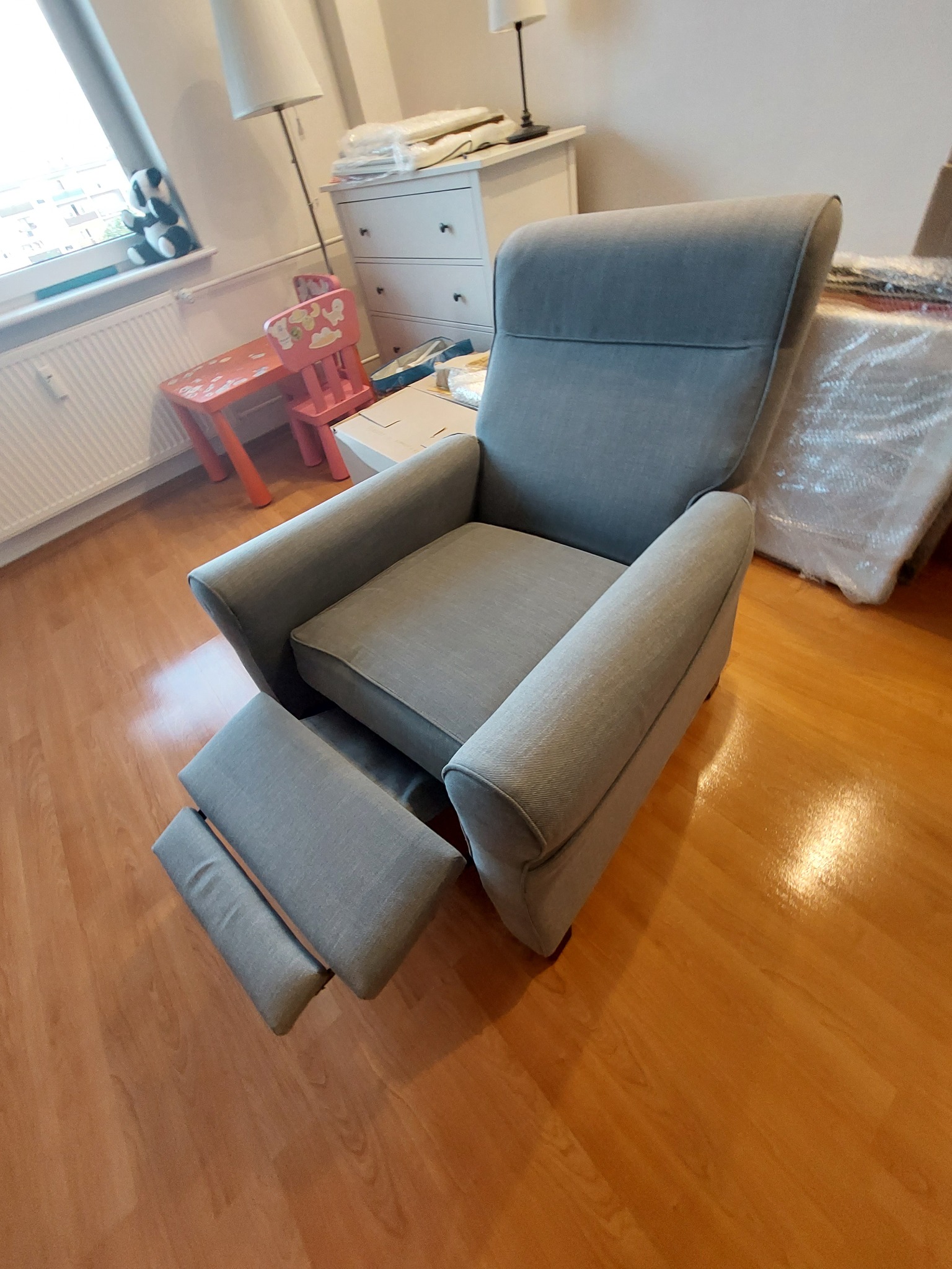 Szary fotel tapicerowany z wysuwanym podnóżkiem na drewnianej podłodze, widok z boku, w tle dziecięcy stolik i krzesełko oraz komoda.