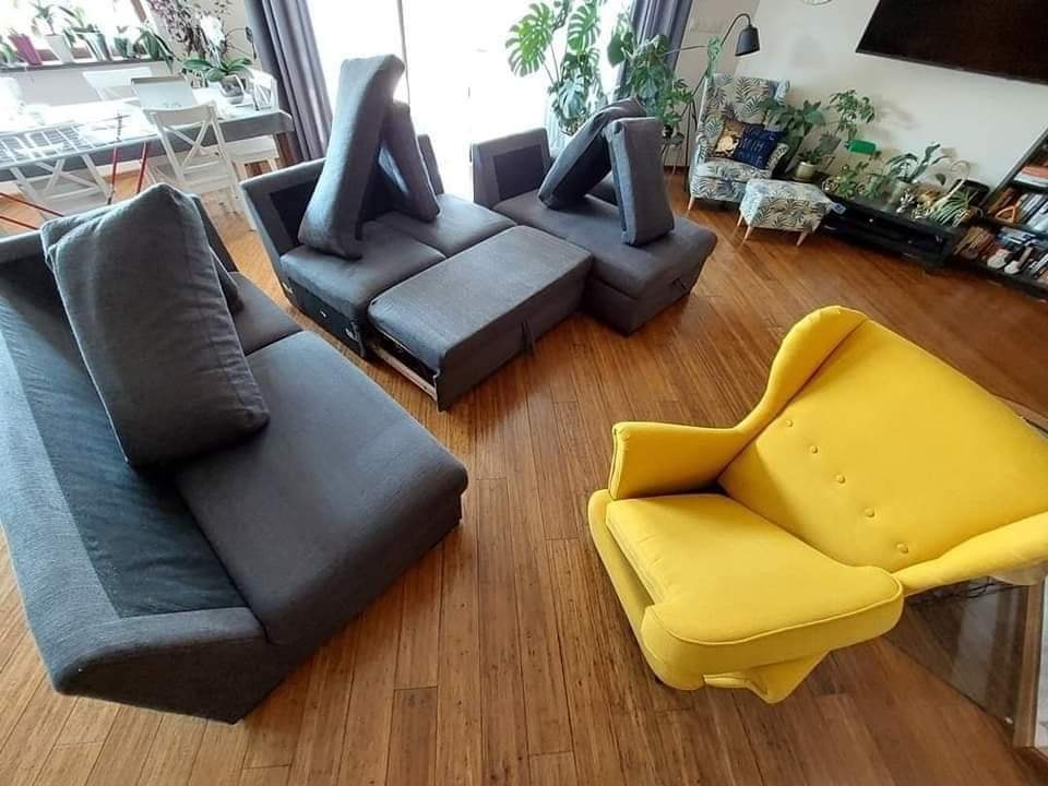 Rozłożona modułowa sofa w odcieniach szarości z podniesionymi oparciami, obok żółty fotel typu uszak na parkiecie, widok z góry, w tle rośliny doniczkowe i stół z krzesłami.