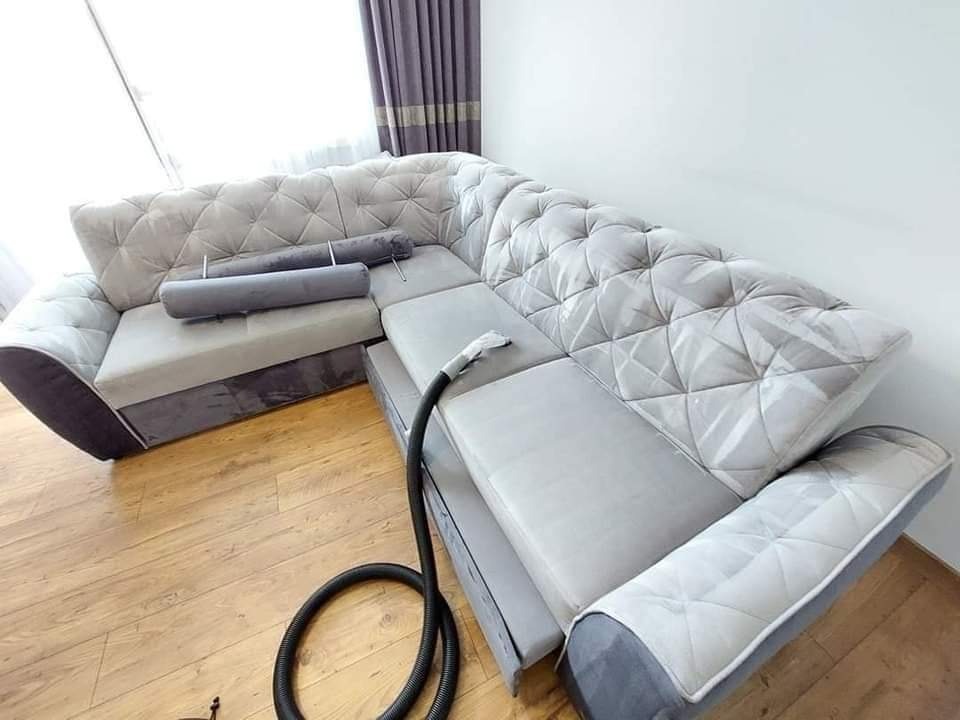 Narożna kanapa z jasną tapicerką czyszczona odkurzaczem piorącym, widoczny wąż odkurzacza na drewnianej podłodze, w tle zasłony i okno.