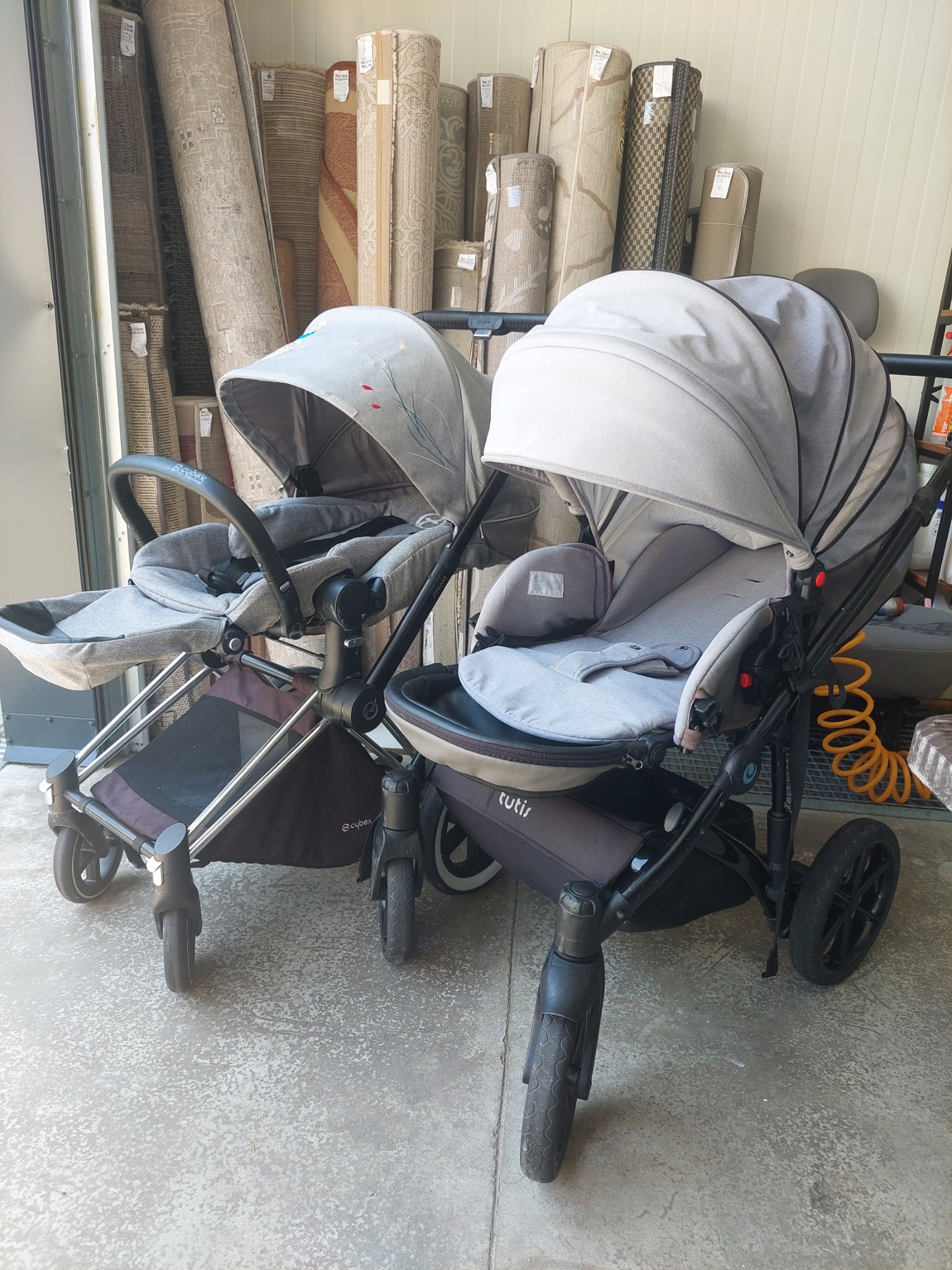 Dwa wózki dziecięce, jeden Cybex, drugi Tutis, stoją na betonowej podłodze na tle zwiniętych dywanów.