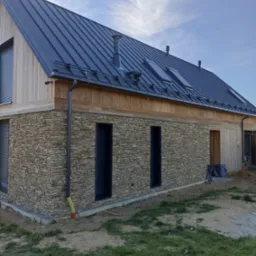 Nowoczesny dom jednorodzinny z ciemną blachą na dachu, drewnianą elewacją i kamienną ścianą, widoczne rynny, okna dachowe i komin.