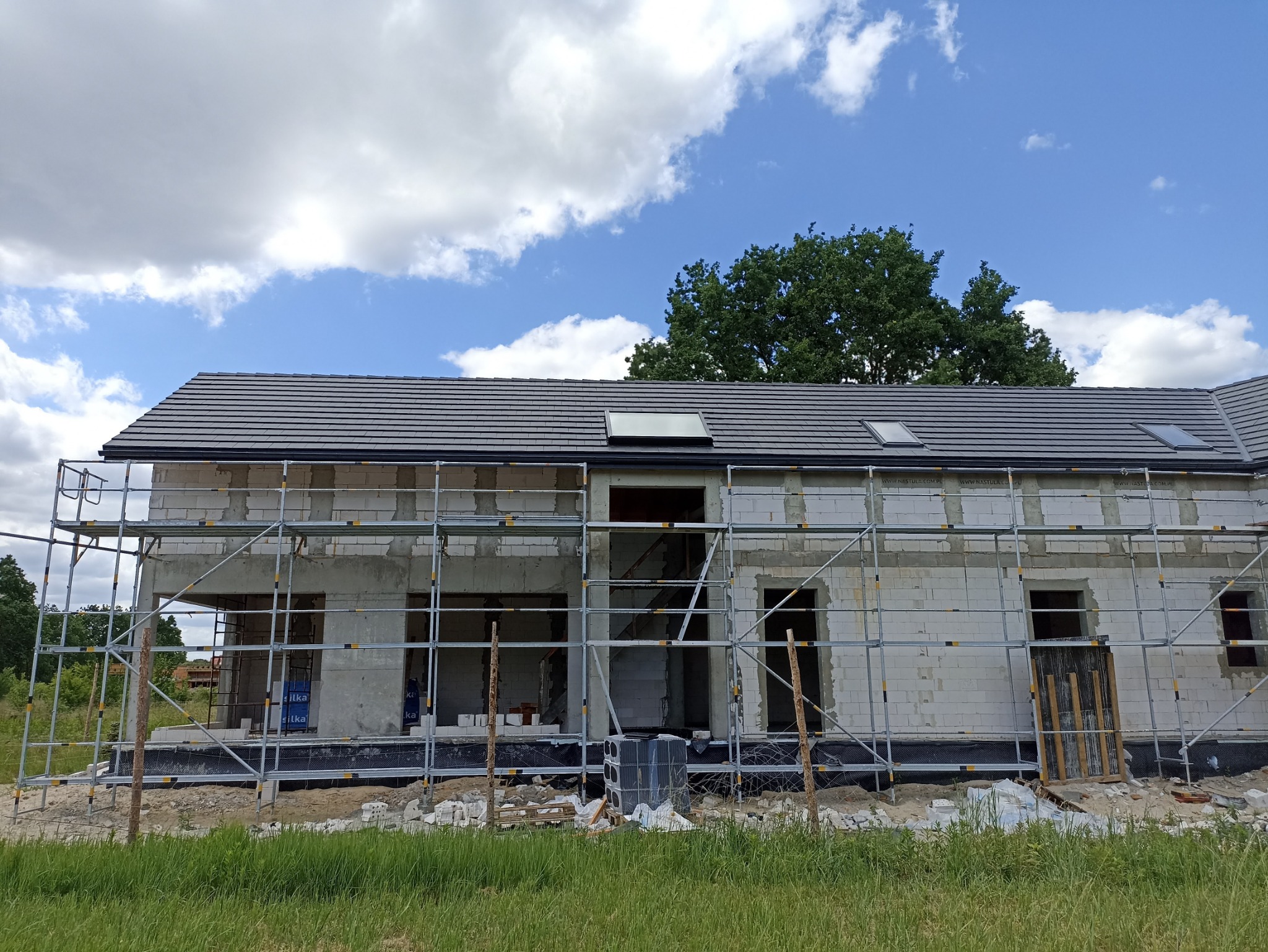 Niedokończony dom z szarym dachem i trzema oknami dachowymi, otoczony rusztowaniem na tle błękitnego nieba z chmurami.