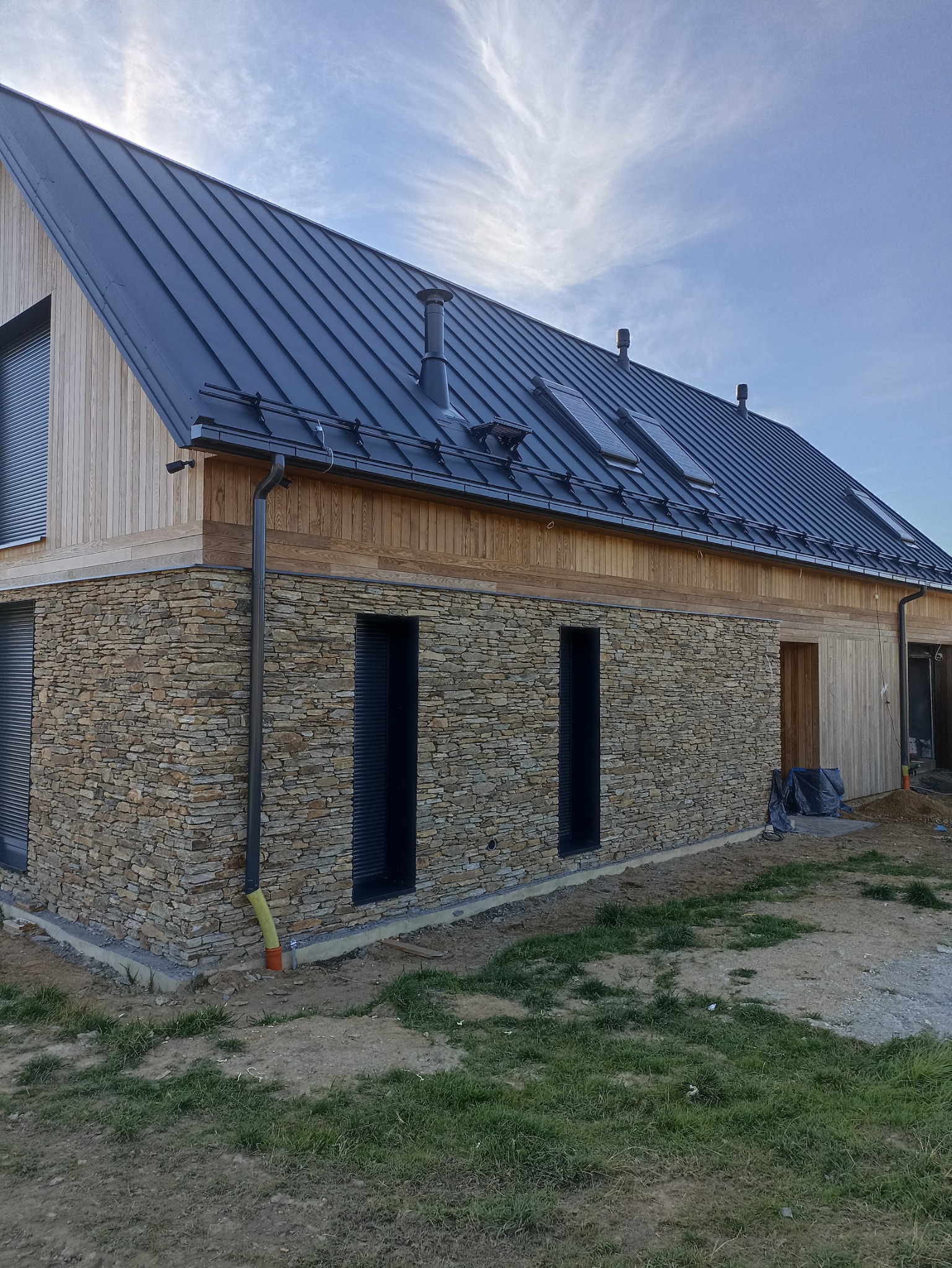 Nowoczesny dom jednorodzinny z ciemną blachą na dachu, drewnianą elewacją i kamienną ścianą, widoczne rynny, okna dachowe i komin.