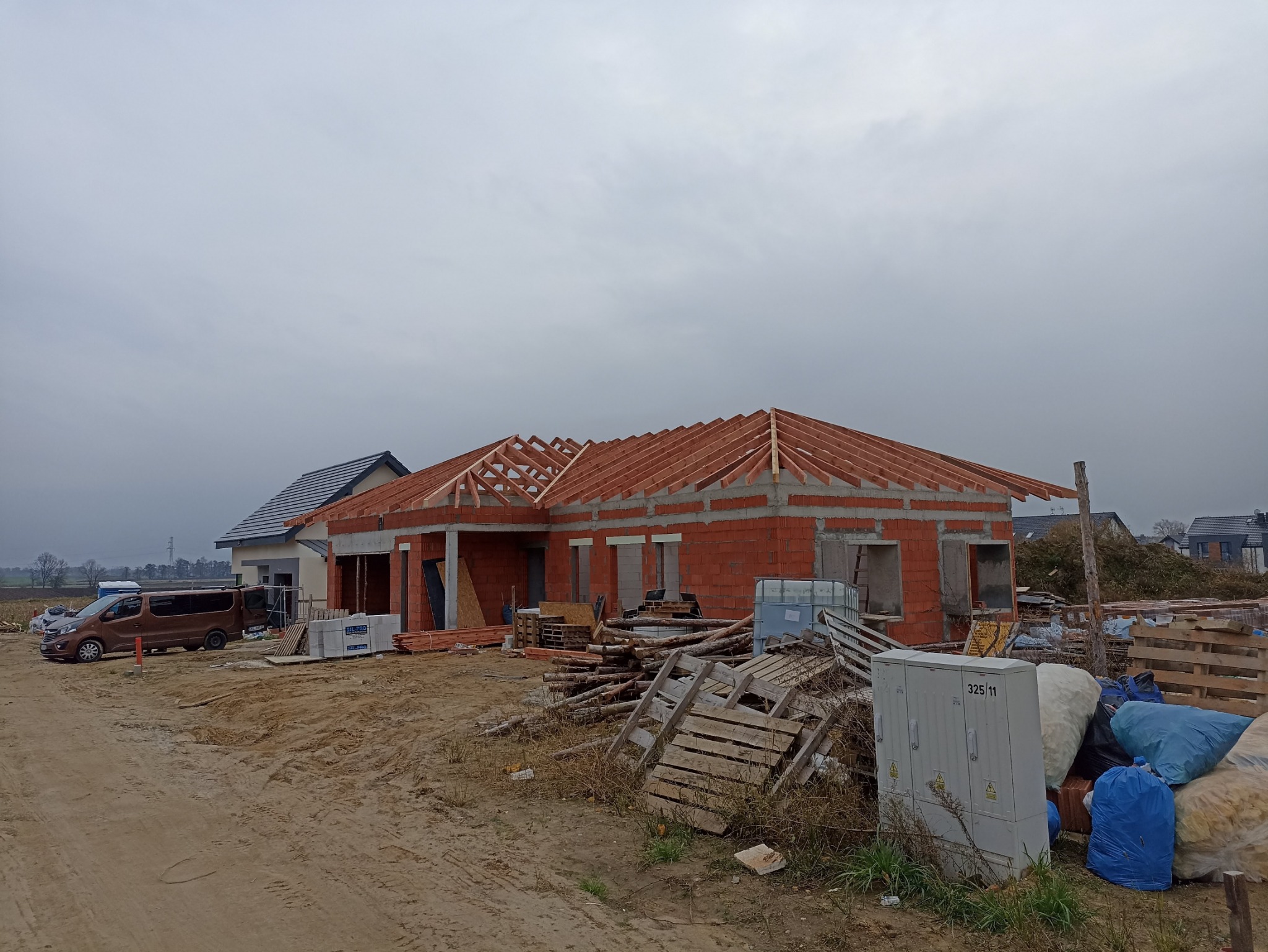 Budowa domu jednorodzinnego w stanie surowym otwartym, widoczna czerwona cegła i drewniana konstrukcja dachu, na pierwszym planie skrzynka elektryczna i gruz budowlany.