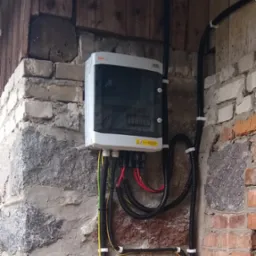 Rozdzielnica elektryczna z przewodami na zewnątrz budynku z kamienno-ceglaną ścianą, widoczne kable w peszlach przymocowane uchwytami do ściany.