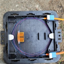 LINK-TOR Sp. z o.o. - Otwarta, czarna skrzynka telekomunikacyjna z włóknami światłowodowymi ułożonymi wewnątrz, pomarańczowe kable z niebieskimi złączkami po bokach, leżąca na betonowym podłożu.