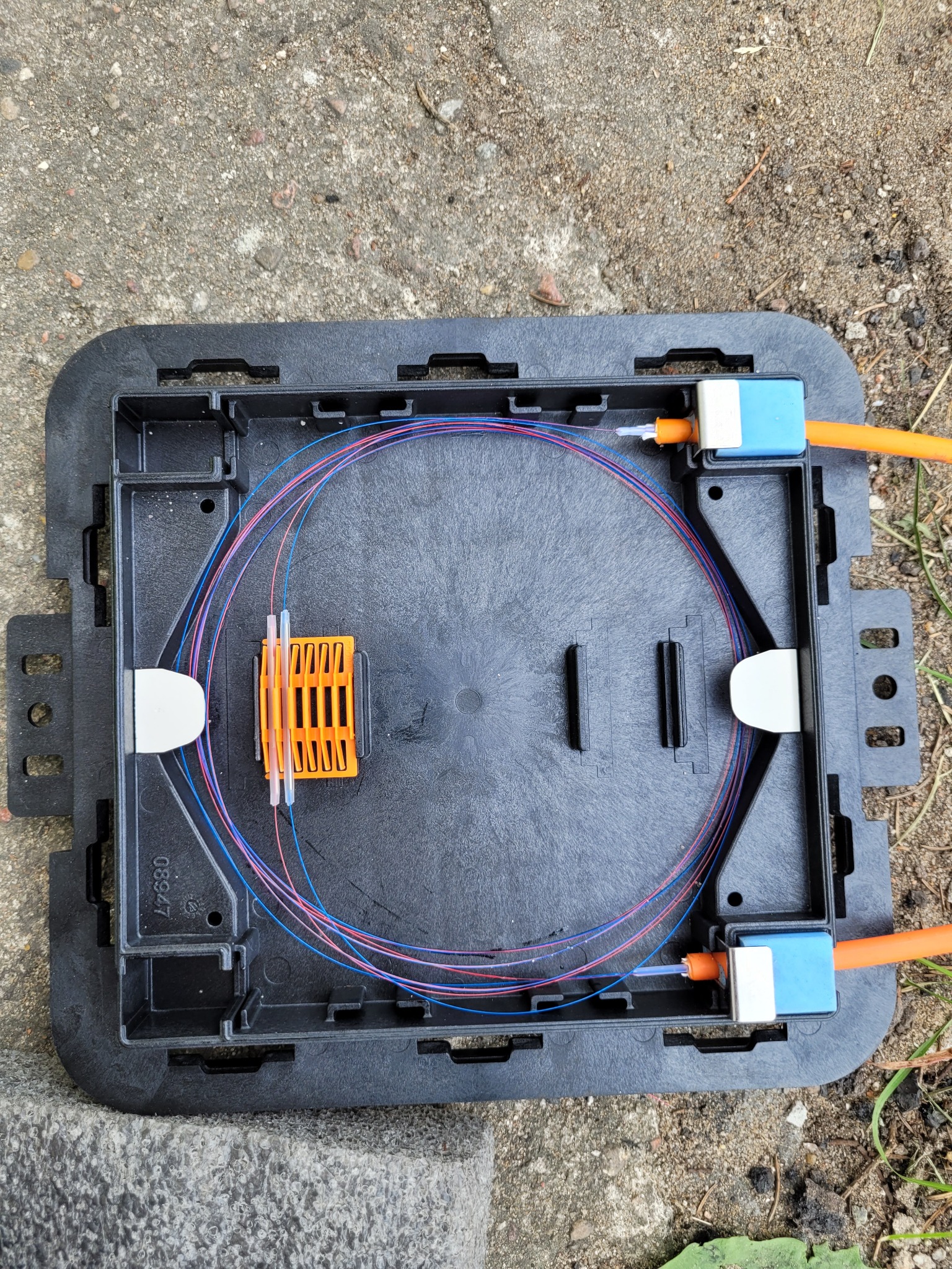 Otwarta, czarna skrzynka telekomunikacyjna z włóknami światłowodowymi ułożonymi wewnątrz, pomarańczowe kable z niebieskimi złączkami po bokach, leżąca na betonowym podłożu.