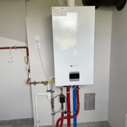 Nowoczesny, biały piec gazowy Saunier Duval zamontowany na ścianie w Tuchowicach, z widocznymi rurami w kolorze czerwonym i niebieskim, oraz czarnym kominem. Szare płytki podłogowe i białe ściany...