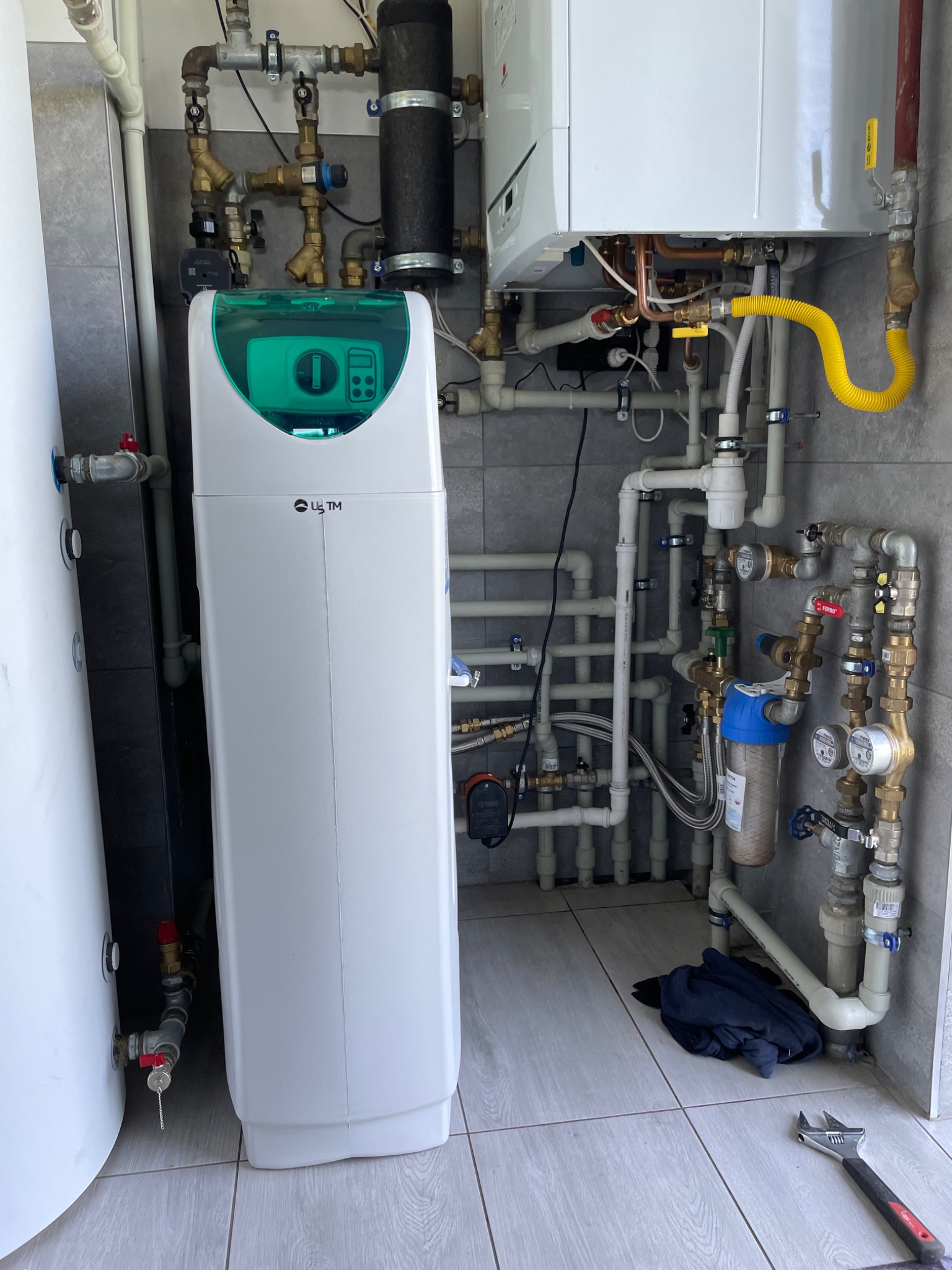 Kompleksowa instalacja hydrauliczna z zmiękczaczem wody, kotłem gazowym i licznymi rurami, widoczny klucz nastawny na podłodze.