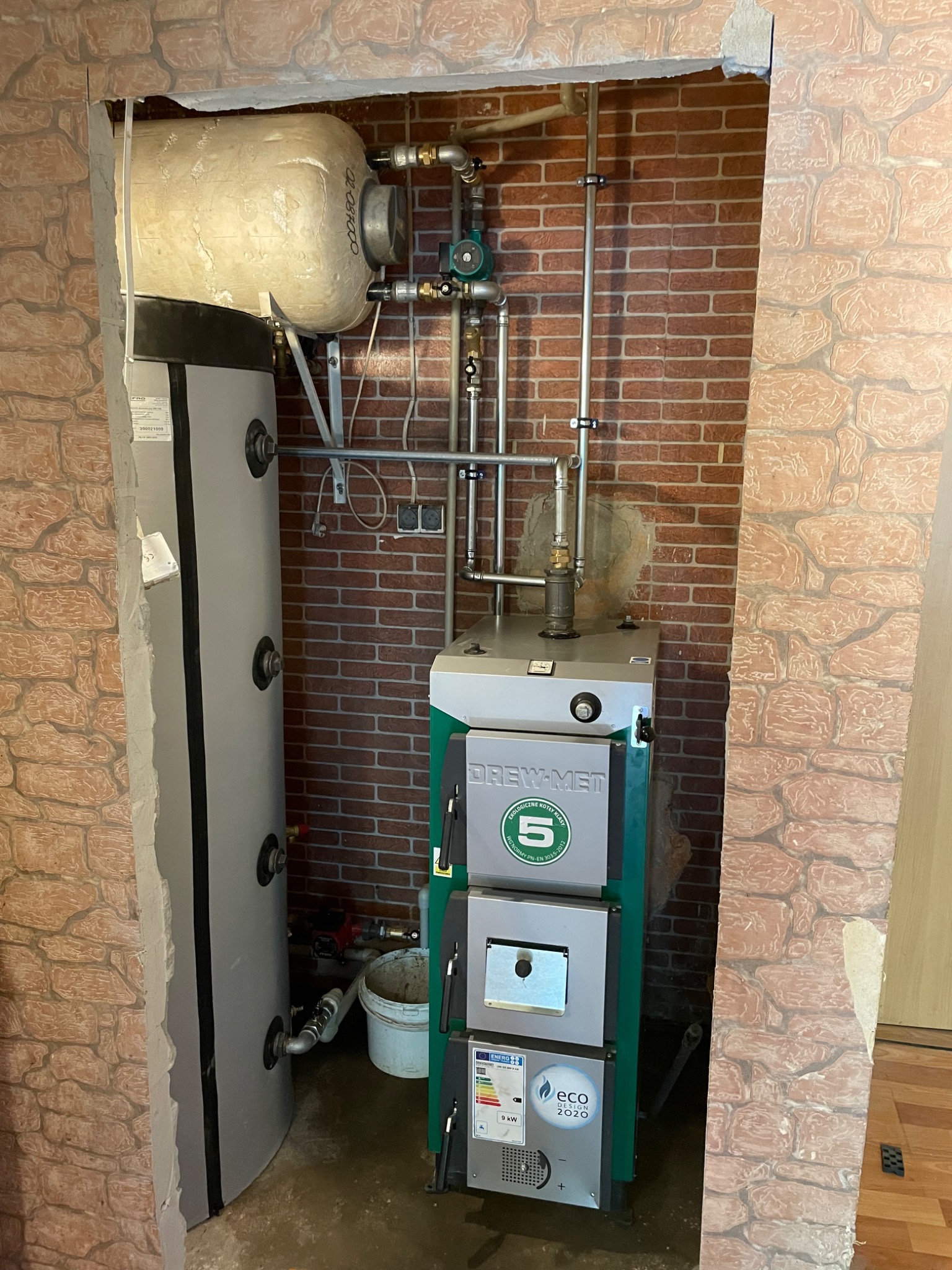 Kotłownia z kotłem Drew-Met Eco Design 2020, zbiornikiem akumulacyjnym i instalacją hydrauliczną na tle ceglanej ściany.