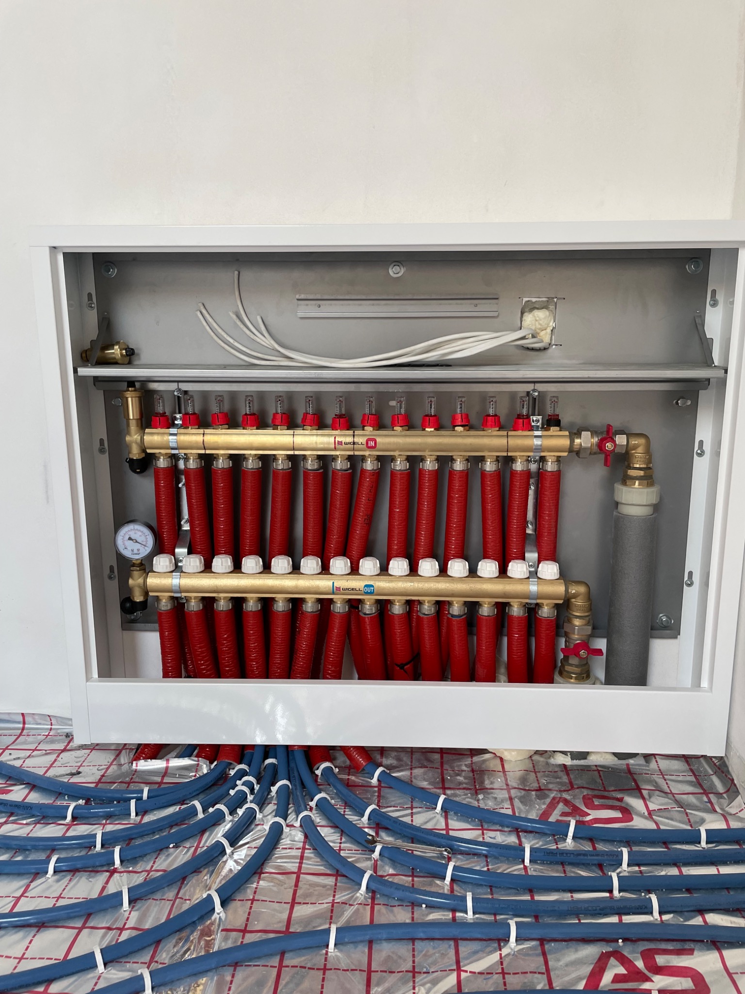 Rozdzielacz ogrzewania podłogowego w białej szafce, widoczne czerwone rury zasilające i niebieskie rury rozprowadzające na folii z siatką, manometr i zawory.