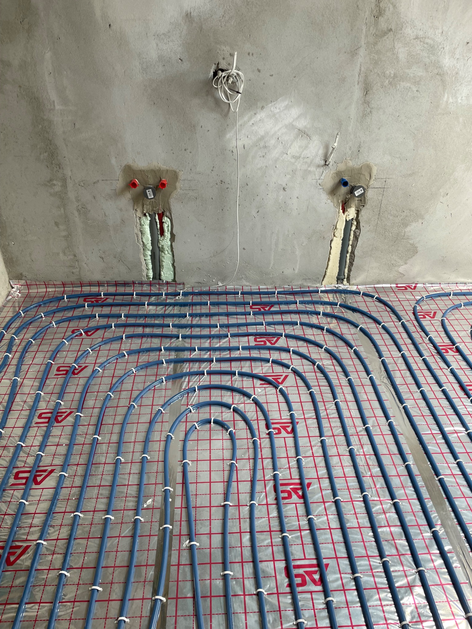 Niedokończona instalacja ogrzewania podłogowego z niebieskich rurek na folii izolacyjnej z czerwonym nadrukiem, widoczne wystające rury ze ściany zabezpieczone pianką montażową.
