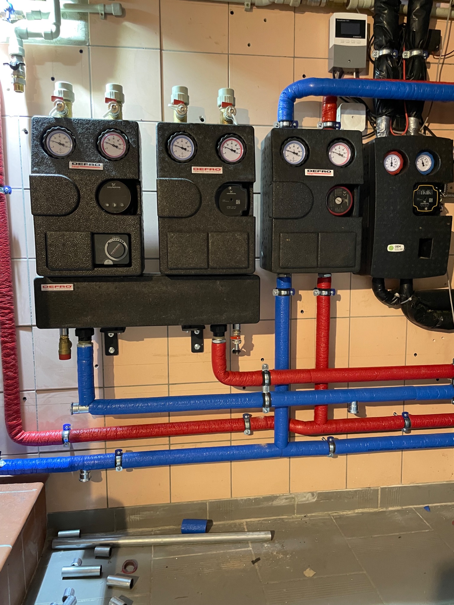 Cztery czarne kotły grzewcze marki Defro zamontowane na kafelkowanej ścianie, z podłączonymi rurami w izolacji koloru niebieskiego i czerwonego. Na podłodze widoczne elementy instalacji.
