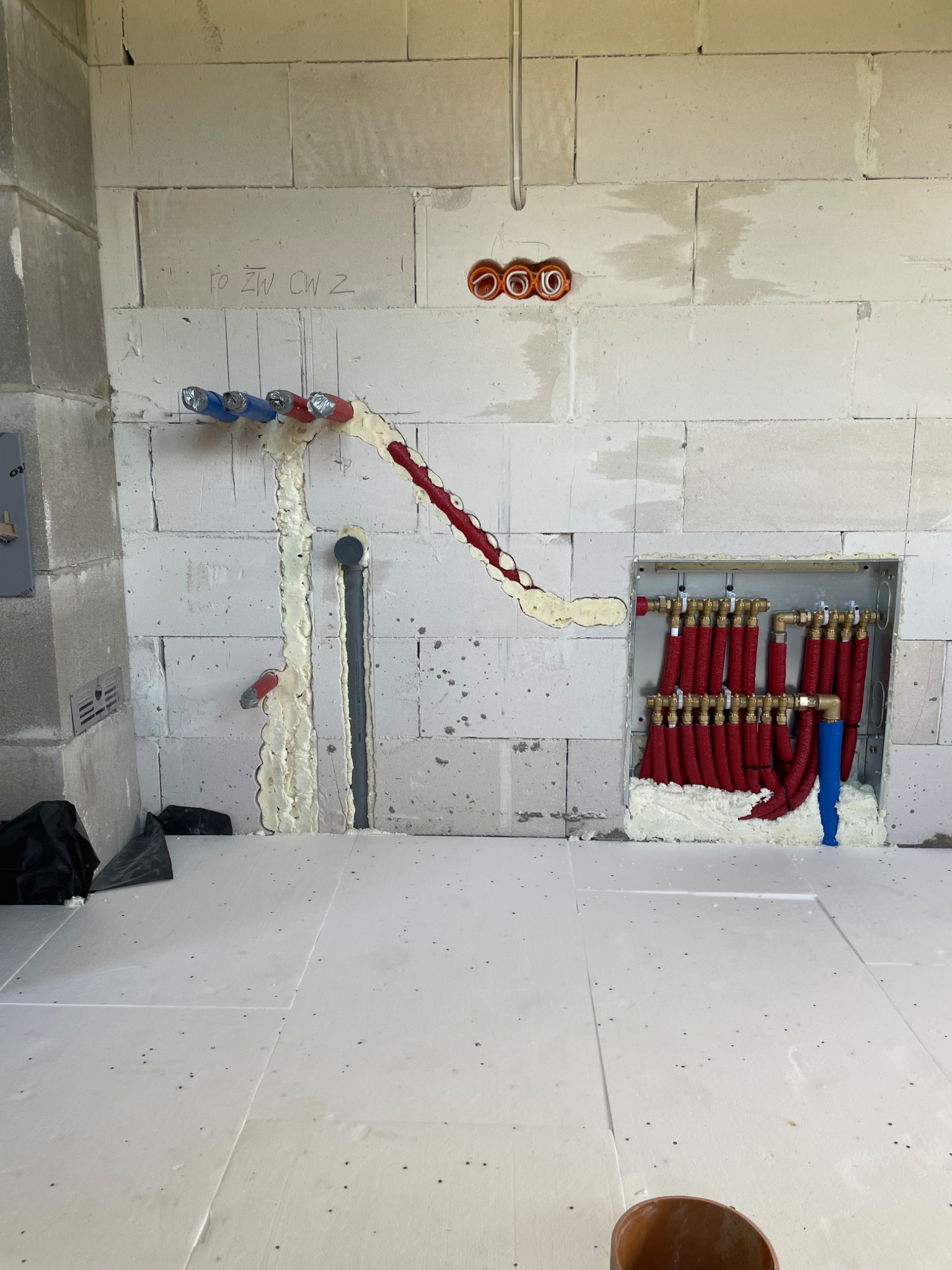 Wnętrze w trakcie prac hydraulicznych: widoczne rury instalacyjne w kolorach czerwonym i niebieskim, zabezpieczone pianką montażową na tle surowych, białych ścian z betonu komórkowego. Część...