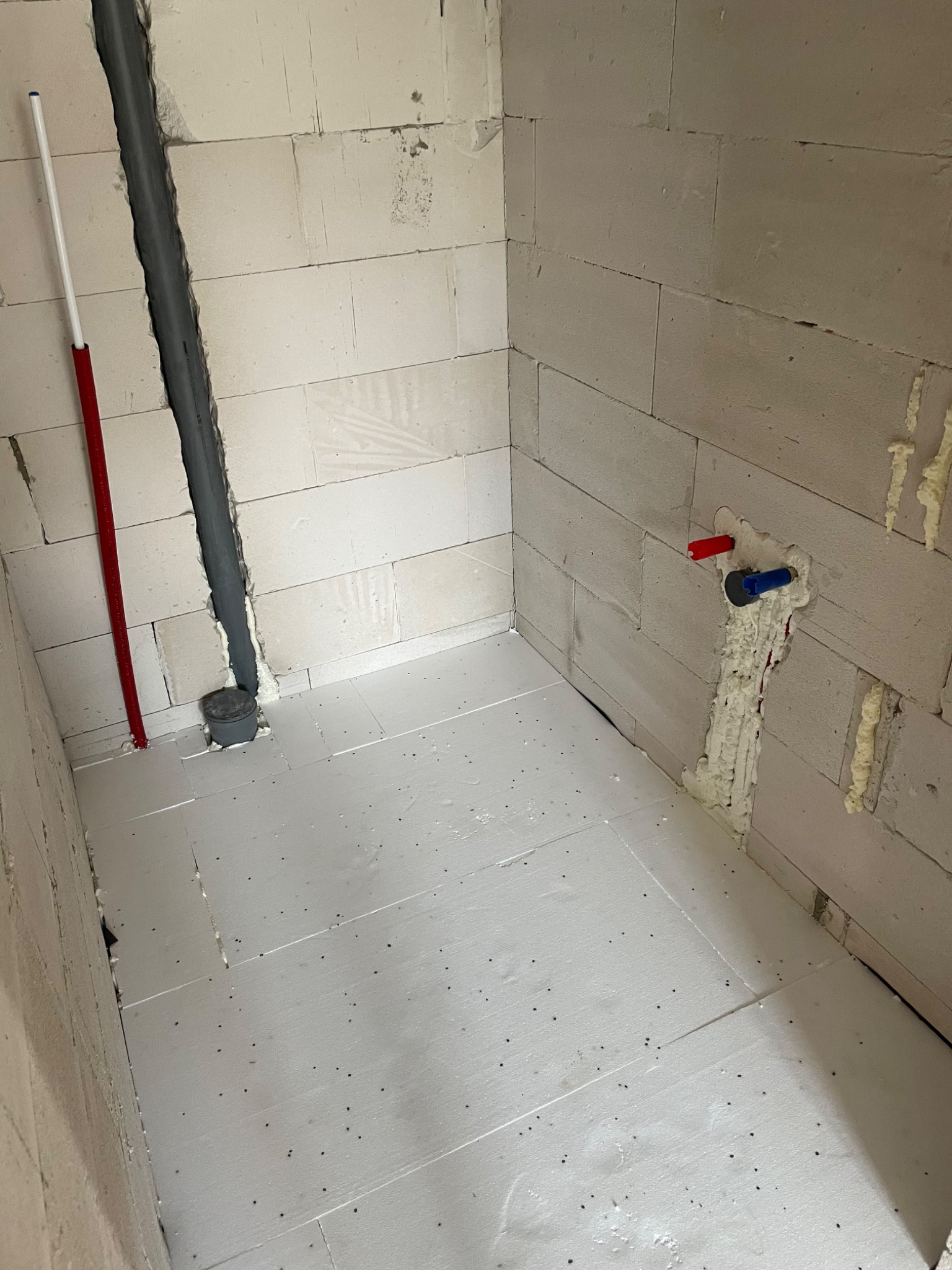 Surowy stan łazienki z widoczną instalacją hydrauliczną w ścianach z betonu komórkowego, rurami grzewczymi i izolacją podłogową ze styropianu.