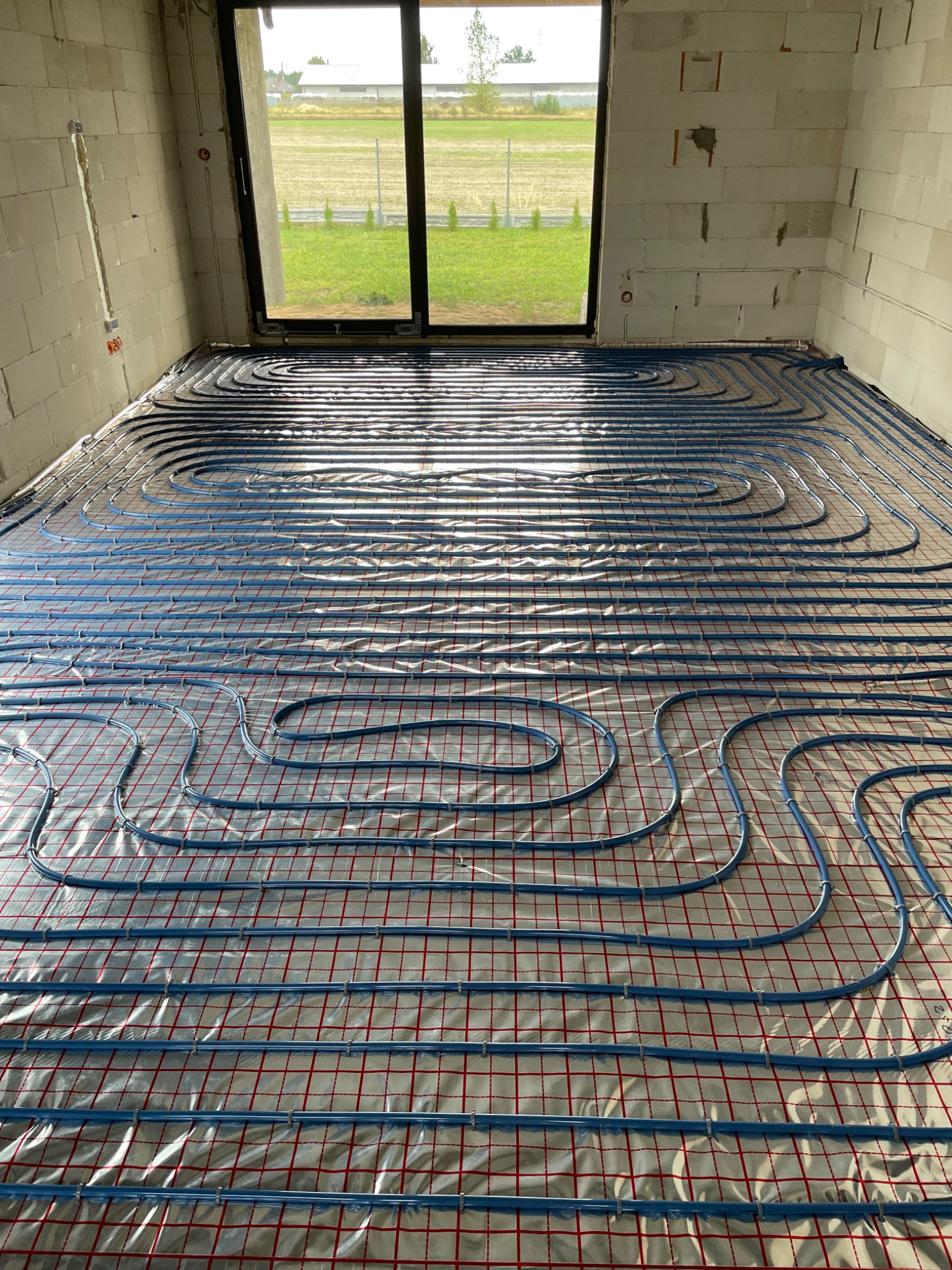 Niebieskie rury ogrzewania podłogowego ułożone na folii z czerwoną siatką w surowym wnętrzu z widokiem na zewnątrz przez okno.