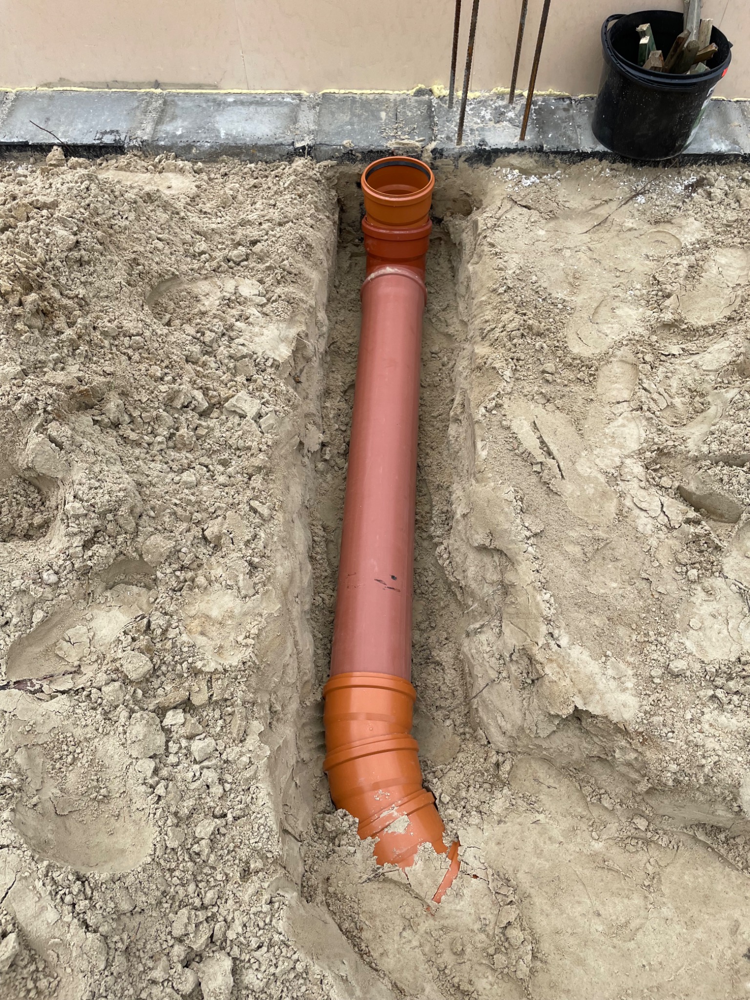 Pomarańczowa rura kanalizacyjna ułożona w wykopie w ziemi, widoczne złącza kielichowe i fragment fundamentu budynku w tle.