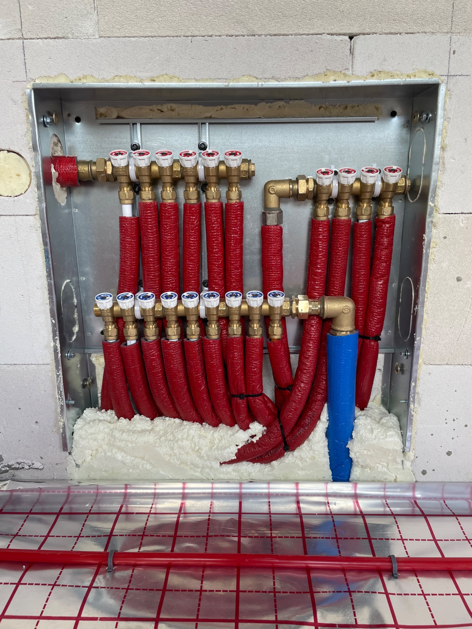 Rozdzielacz ogrzewania podłogowego w metalowej skrzynce, z czerwonymi rurami i białymi zaworami, zamontowany w ścianie z bloczków, pianka montażowa uszczelnia przestrzeń wokół, folia z nadrukiem...