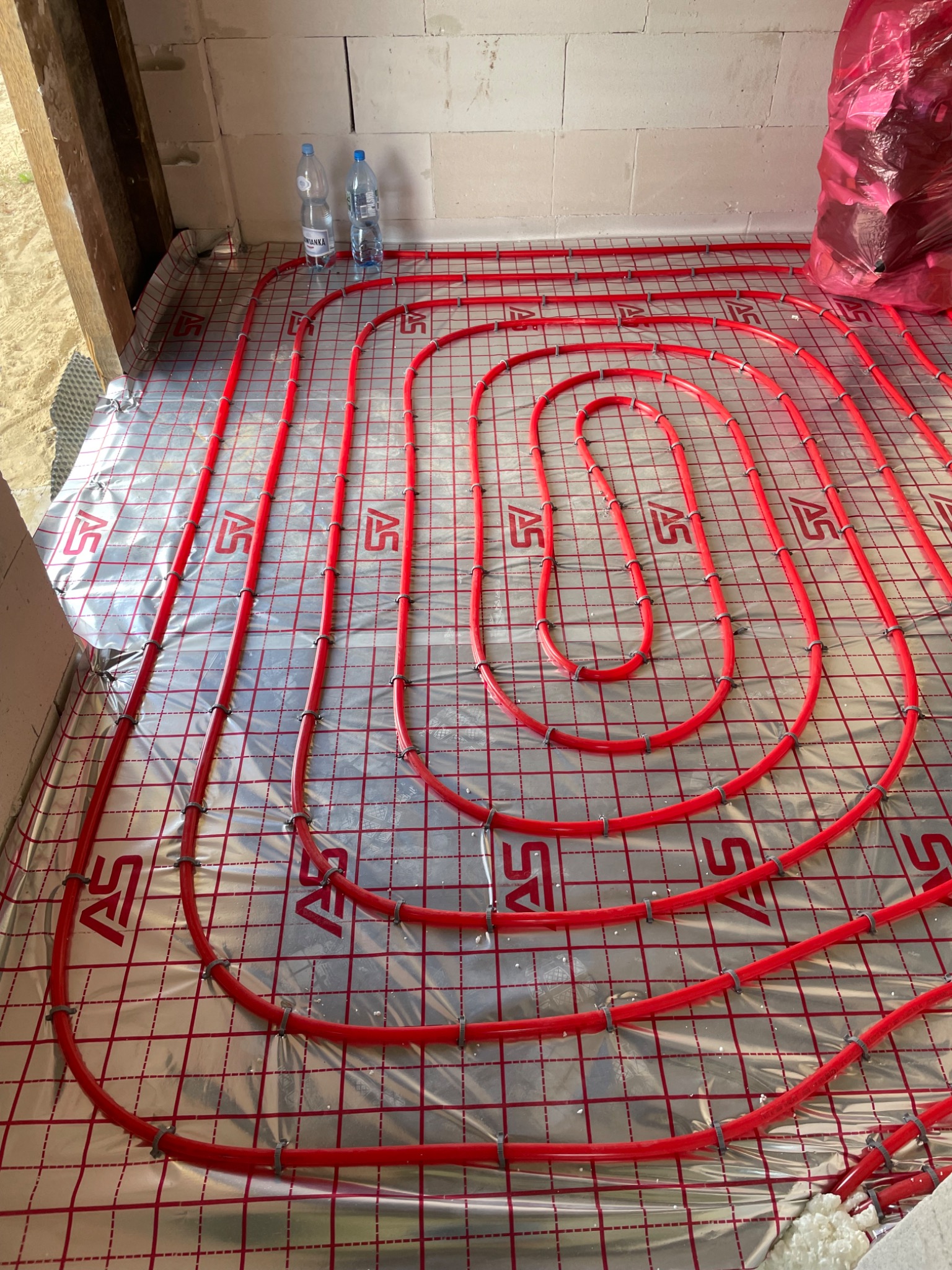 Czerwone rury ogrzewania podłogowego ułożone spiralnie na srebrnej folii z nadrukowaną siatką, zabezpieczone uchwytami, z butelkami wody i workiem w tle.