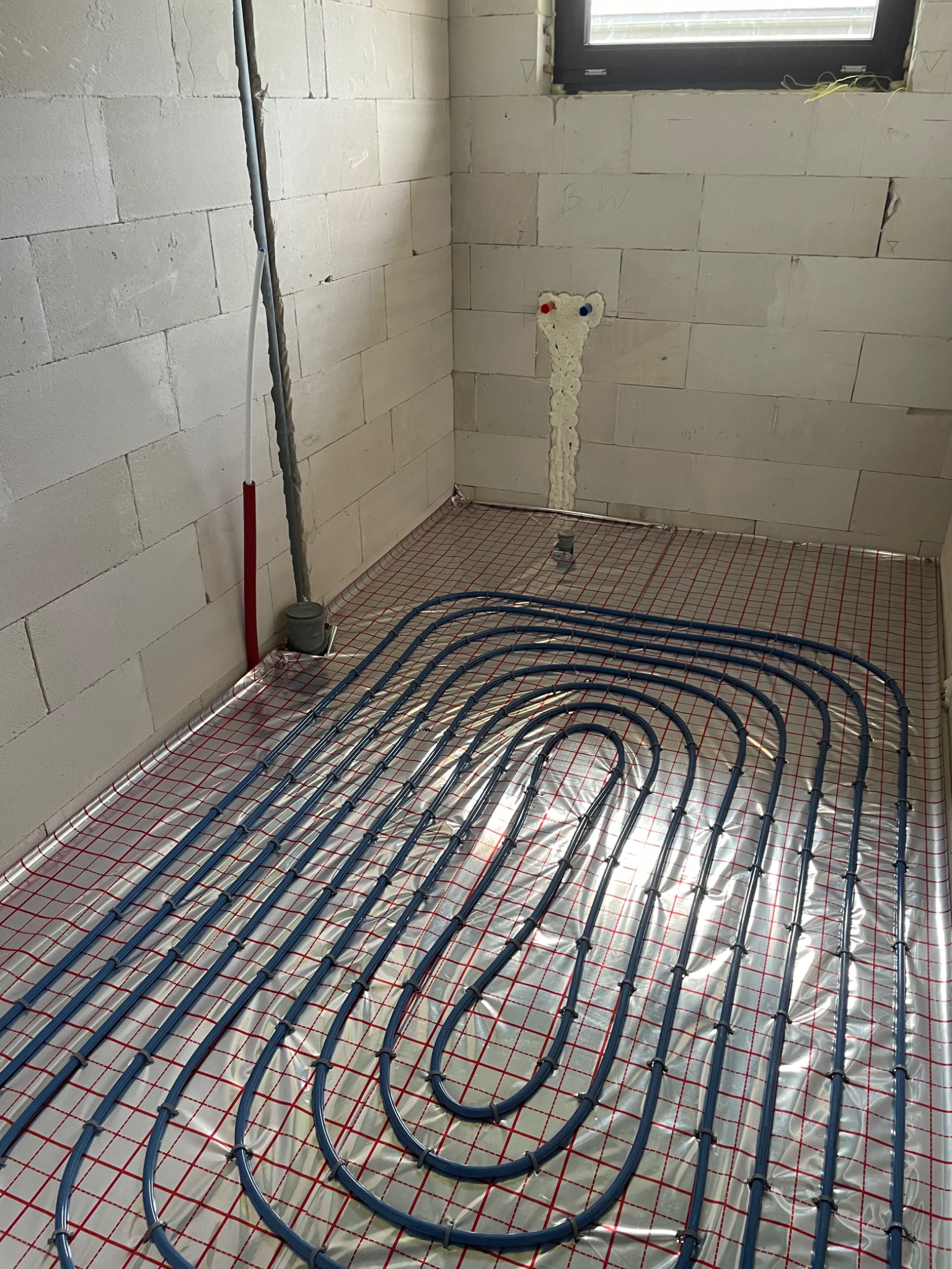 Niebieskie rury ogrzewania podłogowego ułożone na folii z czerwoną siatką w surowym pomieszczeniu z białych bloczków, widoczne wyprowadzenia instalacji wodnej zabezpieczone pianką montażową.