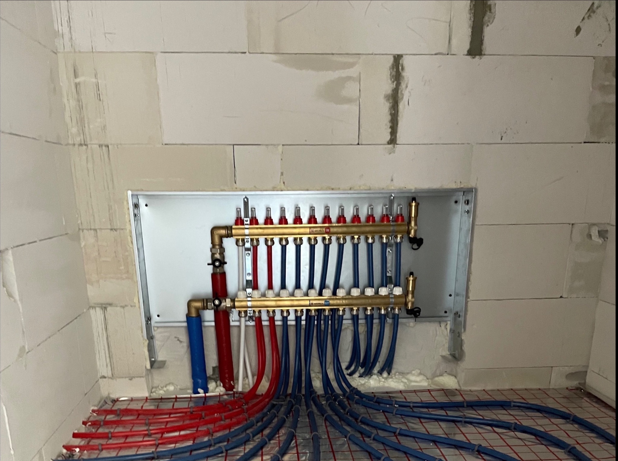 Rozdzielacz ogrzewania podłogowego z rurami w kolorze niebieskim i czerwonym, ułożony na siatce, widoczny na tle ściany z białych bloczków.