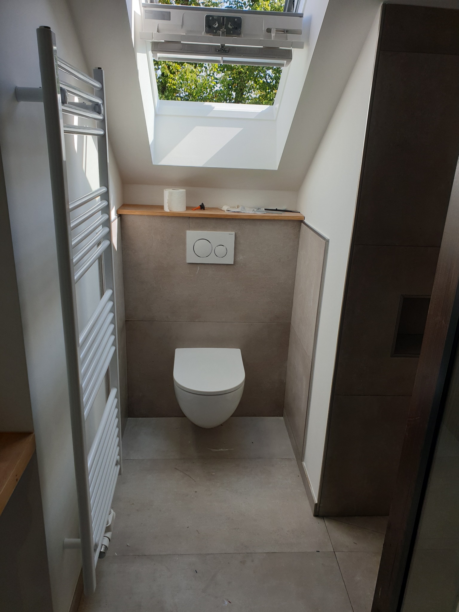 Nowoczesna łazienka na poddaszu z oknem dachowym, wiszącą toaletą i grzejnikiem drabinkowym w kolorze białym, wykończona w szaro-beżowej kolorystyce, częściowo w trakcie prac wykończeniowych...