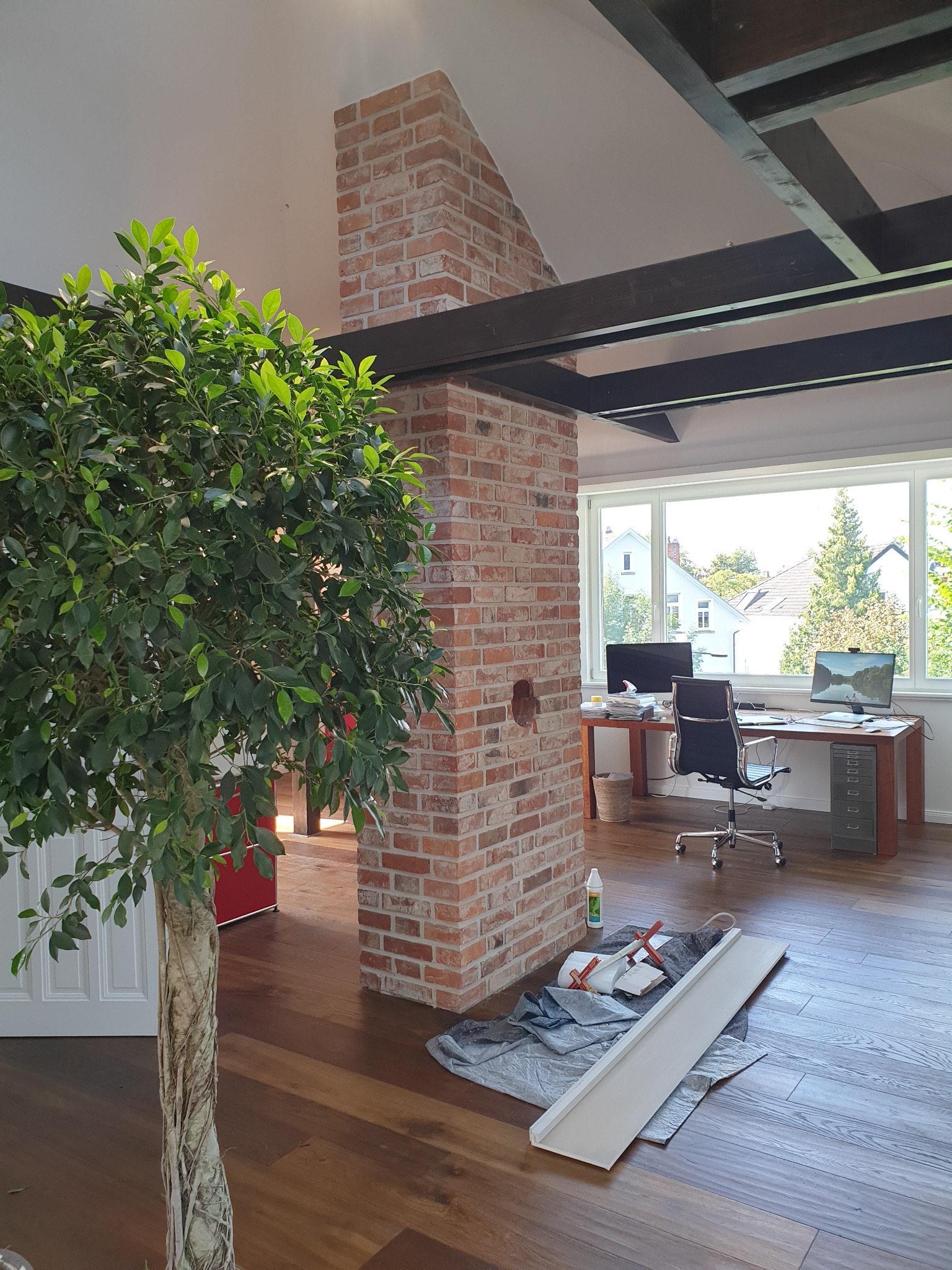Wnętrze z ceglanym filarem i drewnianymi belkami sufitowymi, w tle biurko z komputerami, na podłodze narzędzia i listwa przypodłogowa, w pierwszym planie duża roślina doniczkowa.