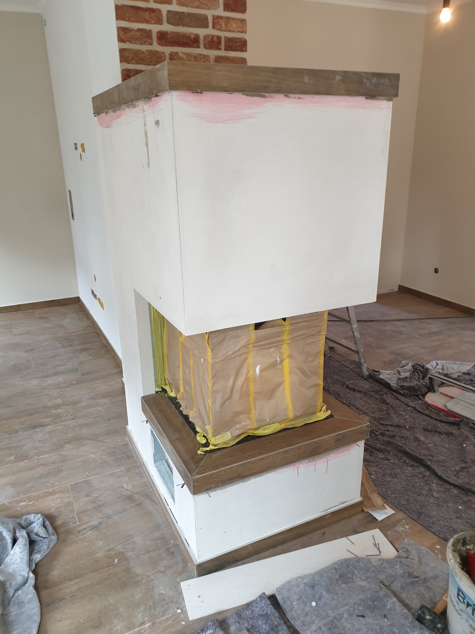 Wnętrze w trakcie prac wykończeniowych: obudowa kominka z białego tynku, zabezpieczona folią i taśmą, z elementami dekoracyjnymi z cegły i drewna, widoczne narzędzia i materiały budowlane na podłodze.