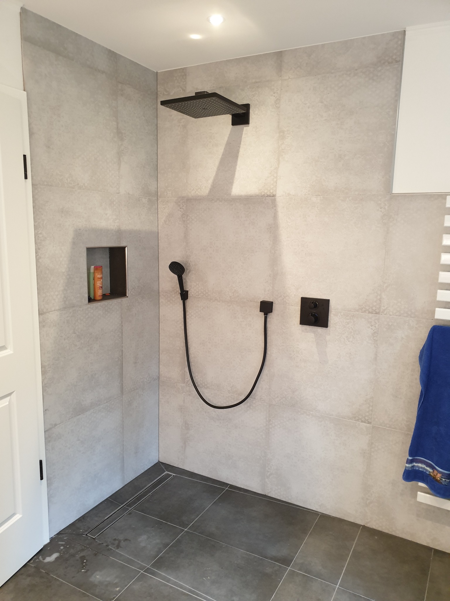 Nowoczesna kabina prysznicowa z szarymi płytkami imitującymi beton, czarną armaturą i wnęką na kosmetyki, widok na odpływ liniowy w podłodze.