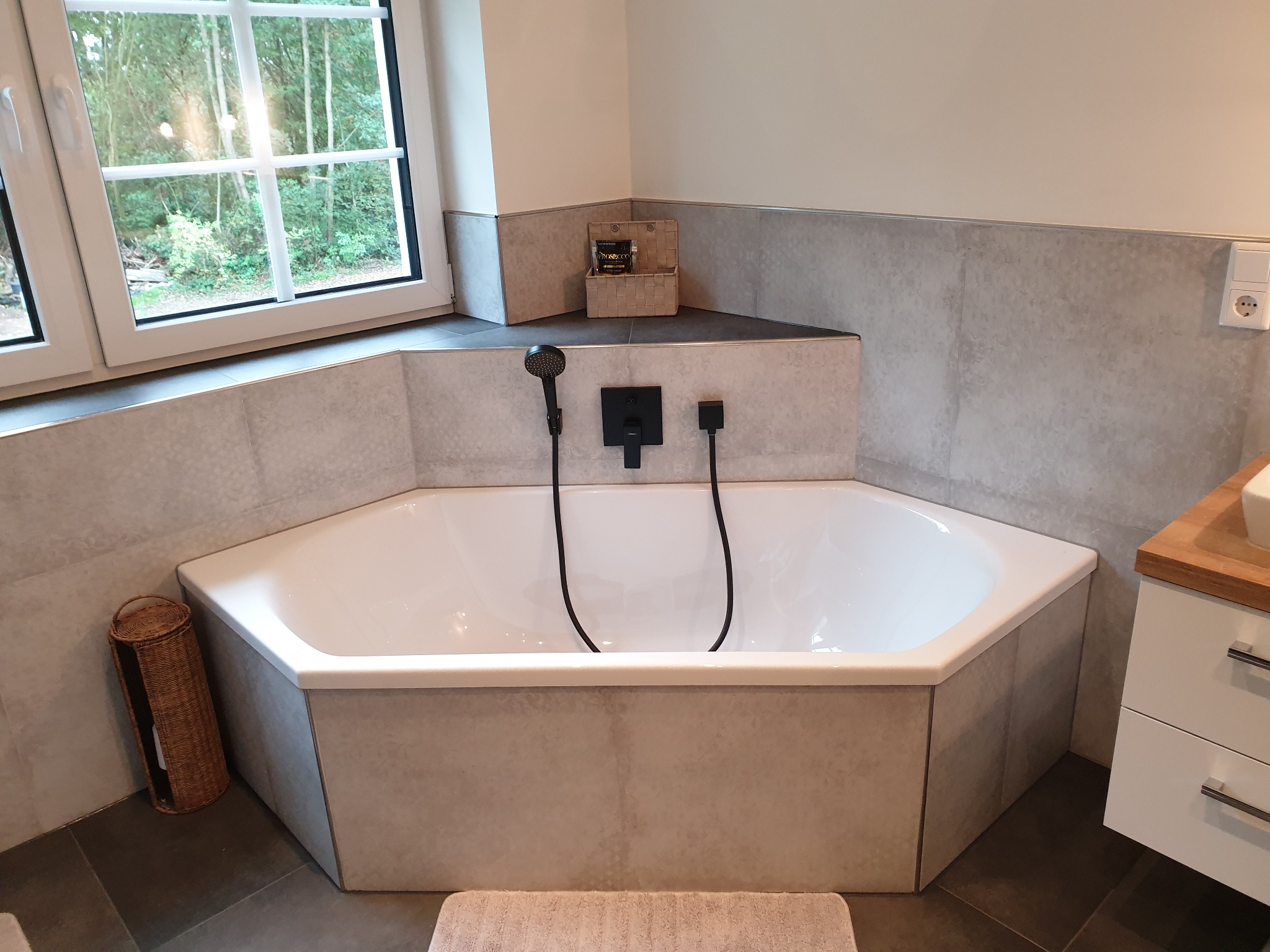 Narożna wanna obudowana płytkami w jasnych odcieniach szarości, z czarną armaturą i widokiem na zieleń za oknem, uzupełnona wiklinowymi akcentami.