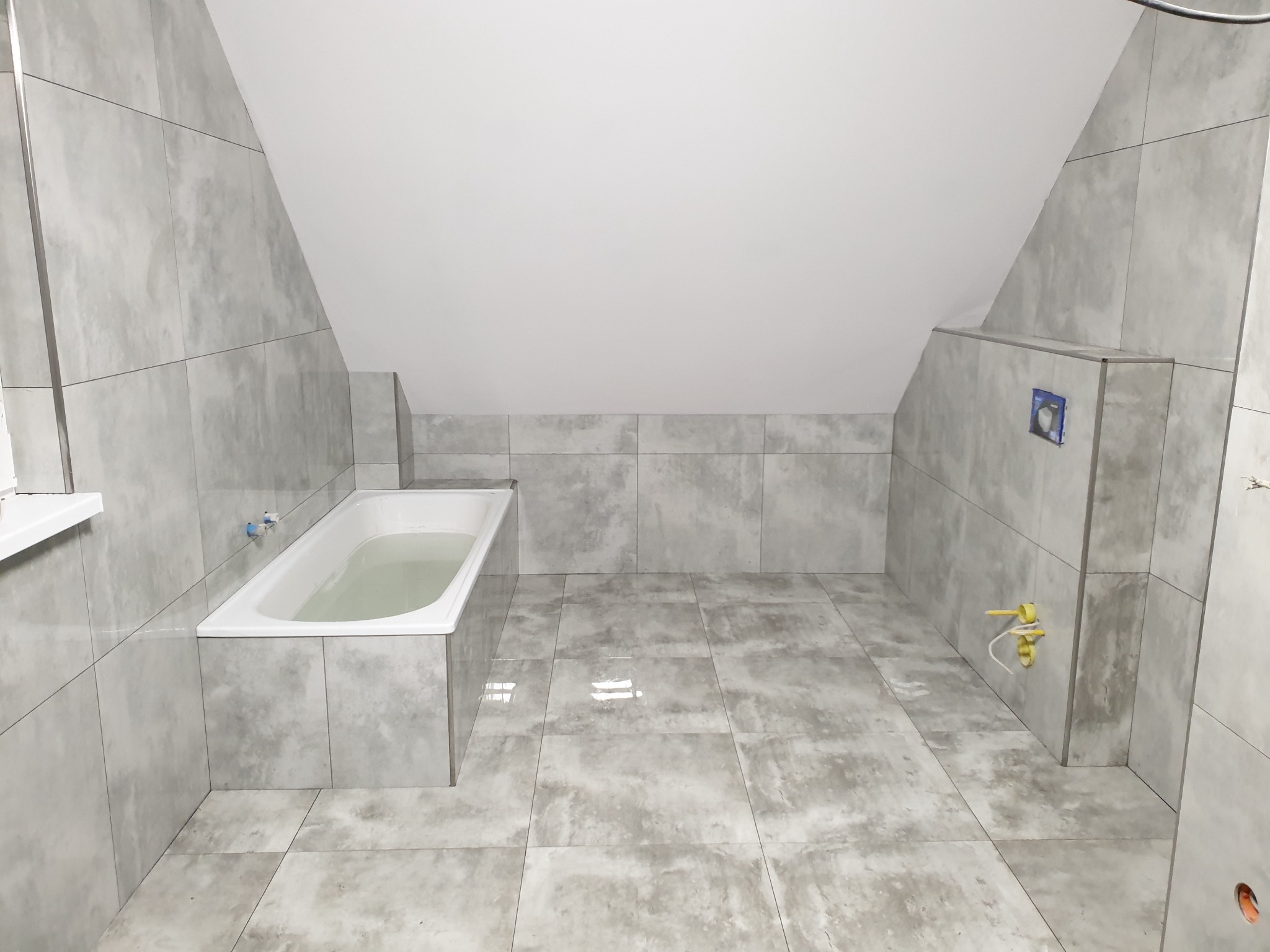 Łazienka na poddaszu ze skośnym sufitem, wykończona szarymi płytkami imitującymi kamień, z białą wanną wypełnioną wodą i widocznymi przyłączami hydraulicznymi przed montażem armatury.